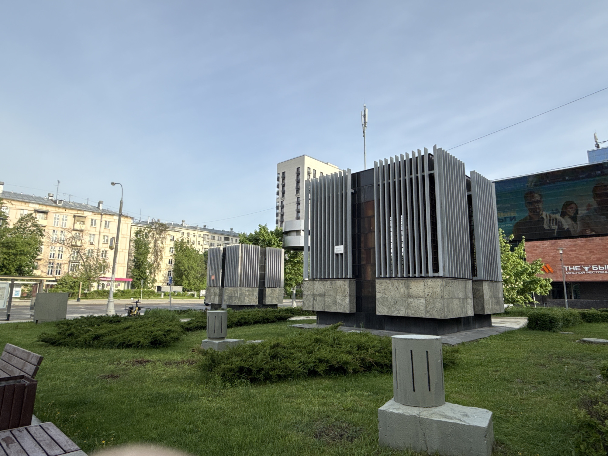 The image shows a monument in Moscow, Russia. It consists of three structures, each composed of vertical metal bars on top of a base of large rectangular stones. In front of the monument are several small cylindrical concrete posts with vertical slits. The monument is situated in a grassy area, surrounded by low bushes. In the background are several buildings, including apartment buildings and a building with a large advertisement. A lamppost is visible in the background, and benches are in the foreground. A pedestrian is riding a bicycle in the distance.