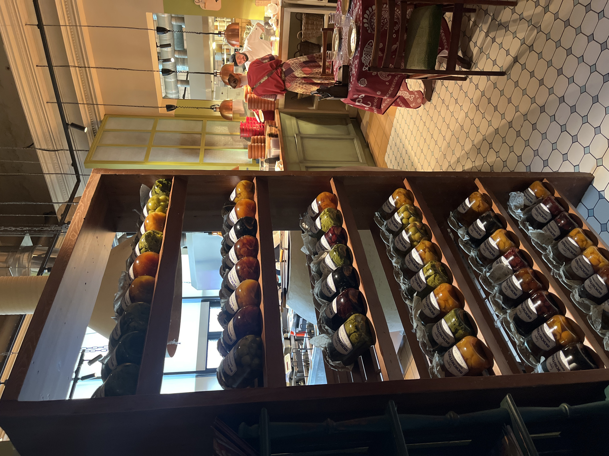 The image shows a rustic wooden shelf filled with jars of preserved food, likely pickled vegetables or fruits. The jars are arranged in rows, with labels on each. The shelf unit is tall and has multiple levels, with the top shelves visible. The background suggests a restaurant or market setting. A woman in a patterned dress is standing at a counter in the background, interacting with a person in a white chef's uniform. There are pendant lights hanging from the ceiling and a tiled floor. A table with a red tablecloth is visible in the foreground. The overall atmosphere is warm and inviting.