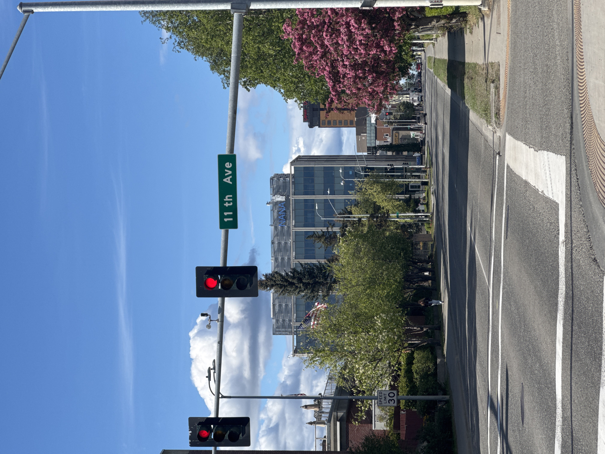The image shows a street scene in Anchorage, Alaska, USA. The foreground is dominated by a paved road with white crosswalk markings. A speed limit sign indicating "30" is visible on the left side of the road. In the middle ground, there are traffic lights showing red, a street sign indicating "11th Ave", trees with green and pink blossoms, and several buildings, including a modern glass-fronted building with "NANA" visible on its facade. An American flag is also visible on a flagpole. The background shows more buildings and trees, with a blue sky and scattered white clouds above. A person is walking on the sidewalk on the right side of the street.