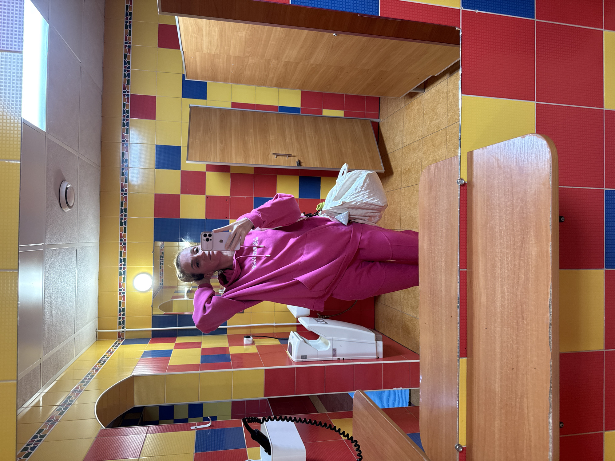 The image shows a woman in a pink tracksuit taking a selfie in a brightly colored public restroom. The restroom walls and floor are tiled in a checkerboard pattern of red, yellow, and blue. There are wooden stall doors on the right side of the frame. A white hand dryer is visible on the wall behind the woman. Above her are wooden cabinets. The environment suggests a public or institutional restroom with a playful, child-friendly design. There is no evidence indicating the restroom's location in Murino, Russia.