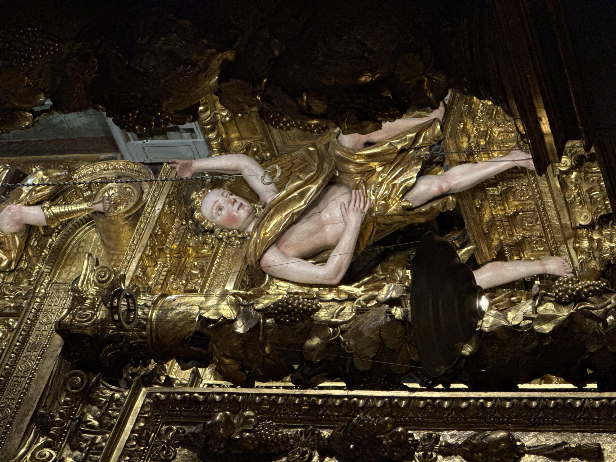This media file captures a gilded baroque sculpture in Santiago de Compostela, Spain. The central subject is a recumbent figure, likely a saint or religious personage, depicted with pale skin, light hair, and rosy cheeks, wearing a golden draped garment. The figure's body is partially exposed, revealing musculature. One arm is extended upwards and bound by a chain to an ornate golden structure above. The other arm rests on the figure's chest.
The sculpture is set within an elaborate, ornate golden altarpiece or retablo, characteristic of Baroque religious art. The golden elements are highly detailed, featuring intricate scrollwork, carved foliage, and clusters of grapes, suggesting themes of fertility, abundance, or possibly the Last Supper. The figure appears to be lying on a raised platform adorned with these decorative motifs. Above the figure, dark, cloud-like or architectural forms are also gilded, creating a dramatic canopy. The lighting suggests an indoor setting, possibly a cathedral or church, with artificial light highlighting the gold and the figure's skin. The overall impression is one of grandeur, opulence, and religious devotion. There is no visible text in the image.