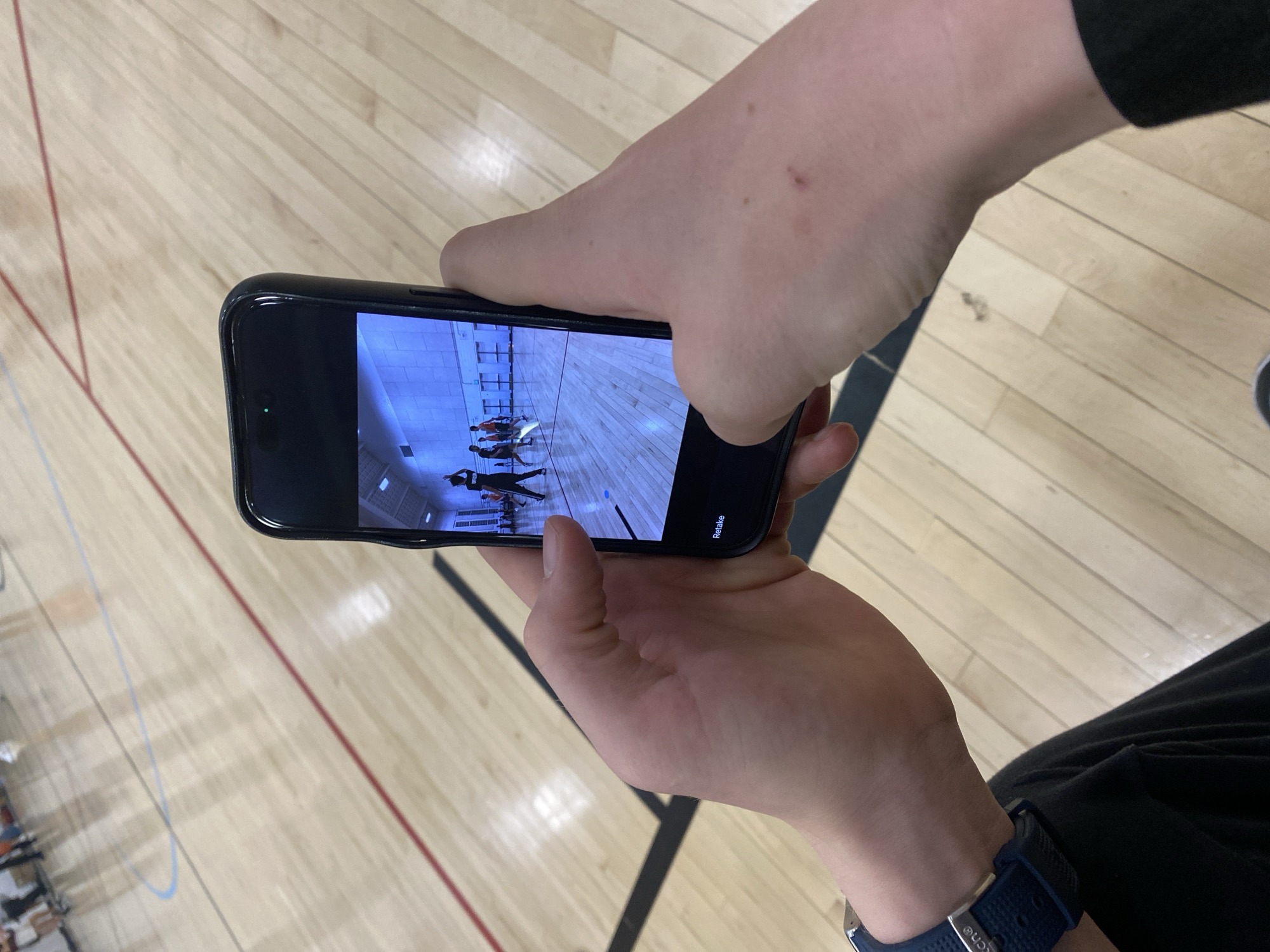 The image depicts a person holding a black smartphone, displaying a photograph of individuals engaged in a dance-like activity in a gymnasium with a wooden floor marked with lines. The person holding the phone has fair skin and is wearing a dark-colored garment and a blue wristwatch on their left wrist. The scene's environment is characterized by the polished wooden floor of the gym, with distinct line markings. The individuals in the photo on the smartphone appear to be wearing athletic attire, engaged in synchronized movement or dance. The location appears to be a gymnasium in a facility within the city of College Park, United States of America.