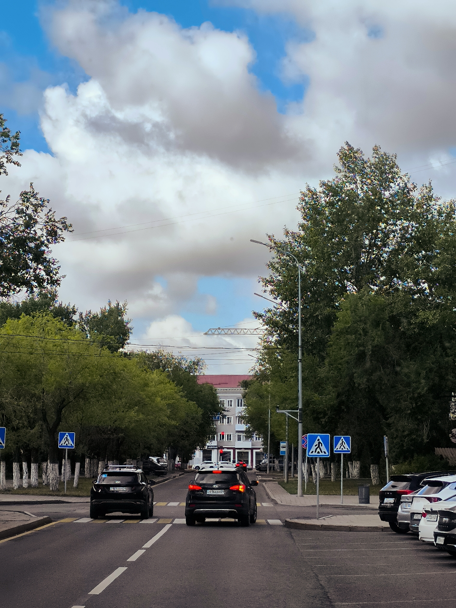 This is a photograph of a street scene in Qarağandı, Kazakhstan. The image is taken from the perspective of a vehicle on the road, looking forward. The sky is partly cloudy with blue patches visible between large white and gray clouds. Large green trees line the sides of the street. In the foreground, there are two black cars on the road, one in each lane, approaching a pedestrian crossing. The car in the right lane has its brake lights on, indicating it is stopping. There are pedestrian crossing signs on poles to the right of the road. Further down the street, more cars are parked. On the left side of the street, there is a sidewalk and a line of white-barked trees. In the distance, buildings are visible, including a multi-story building with a red roof. A construction crane is also visible in the far distance. The overall impression is of a typical urban street on a cloudy day.