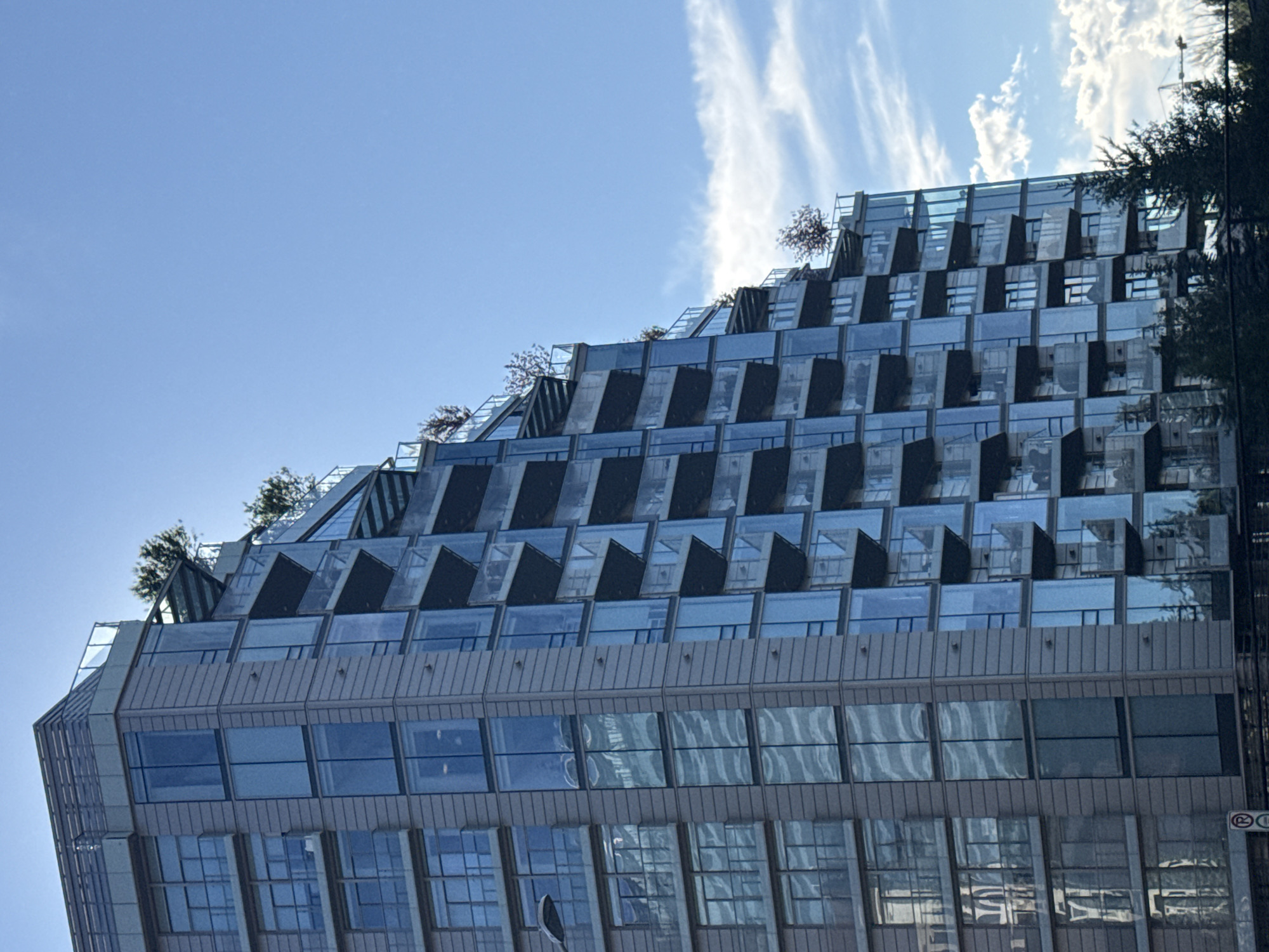 This is an exterior, eye-level, medium shot of a modern, stepped, high-rise residential building with a glass facade and balconies, located in Calgary, Canada. The building is tall and angular, with the upper floors set back from the lower floors, creating a terraced effect. Several trees are visible on the uppermost balconies. The sky is blue with scattered white clouds. The building's architecture suggests it is a contemporary condominium or apartment complex. The lower portion of the image shows some dark green foliage, likely trees or bushes, and a portion of a dark horizontal structure that could be a fence or barrier. A circular red and white sign, possibly a traffic sign, is partially visible on the far left of the image.