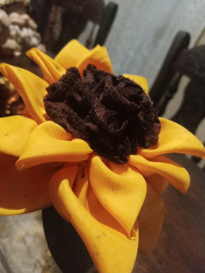 A crafted sunflower dominates the frame, showcasing bright yellow petals made of fabric. The petals are arranged in layers, some folded or curved to add depth. At the flower's center sits a textured, dark brown mass resembling seeds. The sunflower appears to be resting on a surface, with background elements including hints of furniture and other objects, all out of focus. The lighting suggests an indoor setting, casting shadows on the flower's petals.