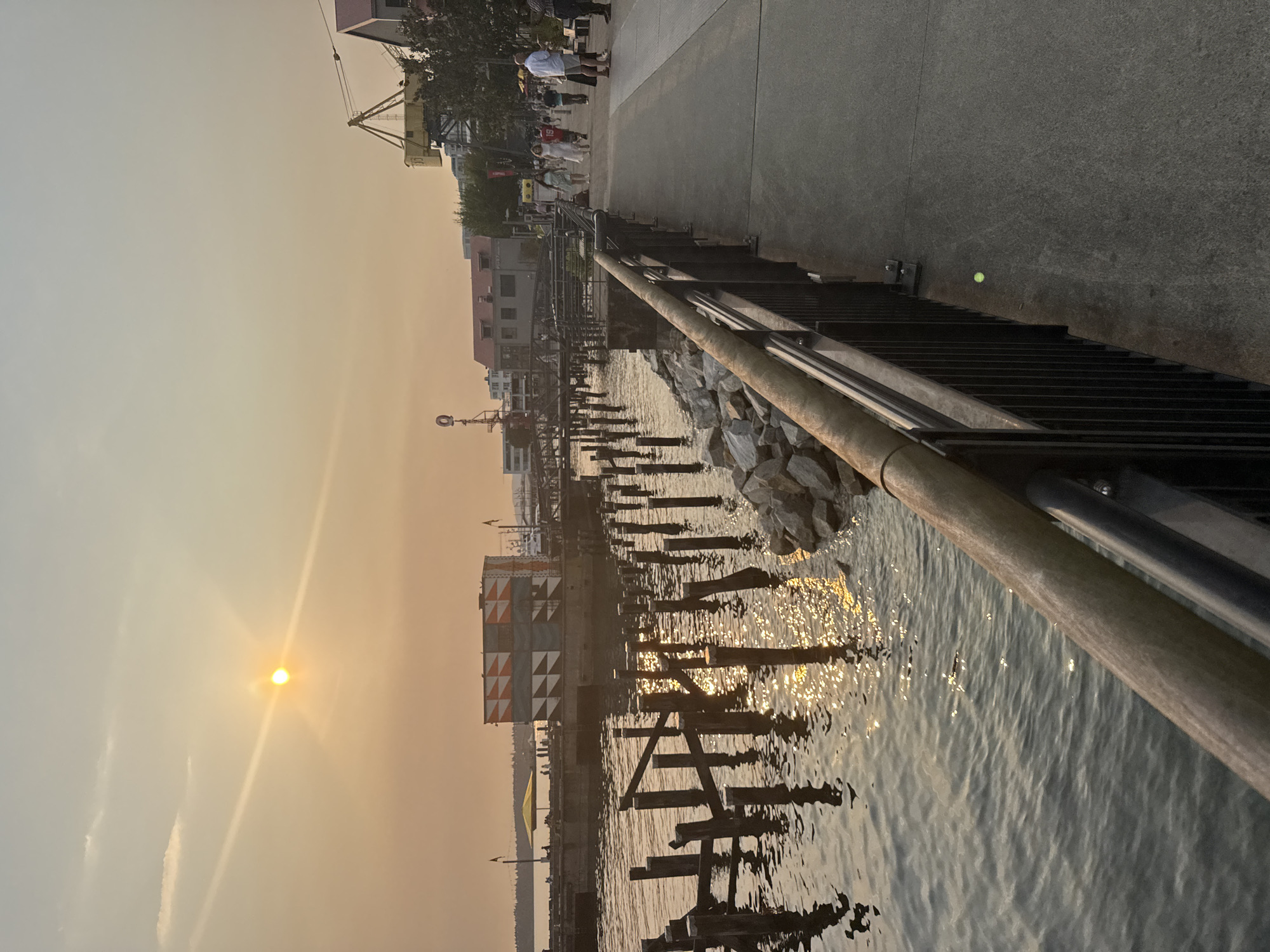The image shows a waterfront promenade in North Vancouver, Canada, likely at Lonsdale Quay during sunset. The sun, a hazy orange orb, is visible in the upper left quadrant of the sky, casting a warm, diffused light over the scene. Smoke in the atmosphere contributes to the orange hue of the sky and the soft lighting.
On the left side of the image, the water is choppy, reflecting the golden light of the setting sun. Numerous old wooden pilings, remnants of a former pier or dock, jut out from the water, some broken and leaning. A section of rocky shoreline is visible in the foreground.
A paved walkway runs along the right side of the image, bordered by a black metal railing with a wooden handrail. Several people are strolling along the walkway, dressed in casual attire. They appear to be enjoying the evening.
In the mid-ground, a modern, box-like structure with a geometric pattern of orange and white on its sides is situated on a pier. Beyond that, buildings with red roofs and industrial cranes are visible, suggesting a port or commercial area. A yellow umbrella is set up on the pier near the modern structure, providing shade for an unseen area. The overall atmosphere is one of a relaxed evening at a waterfront urban setting.
