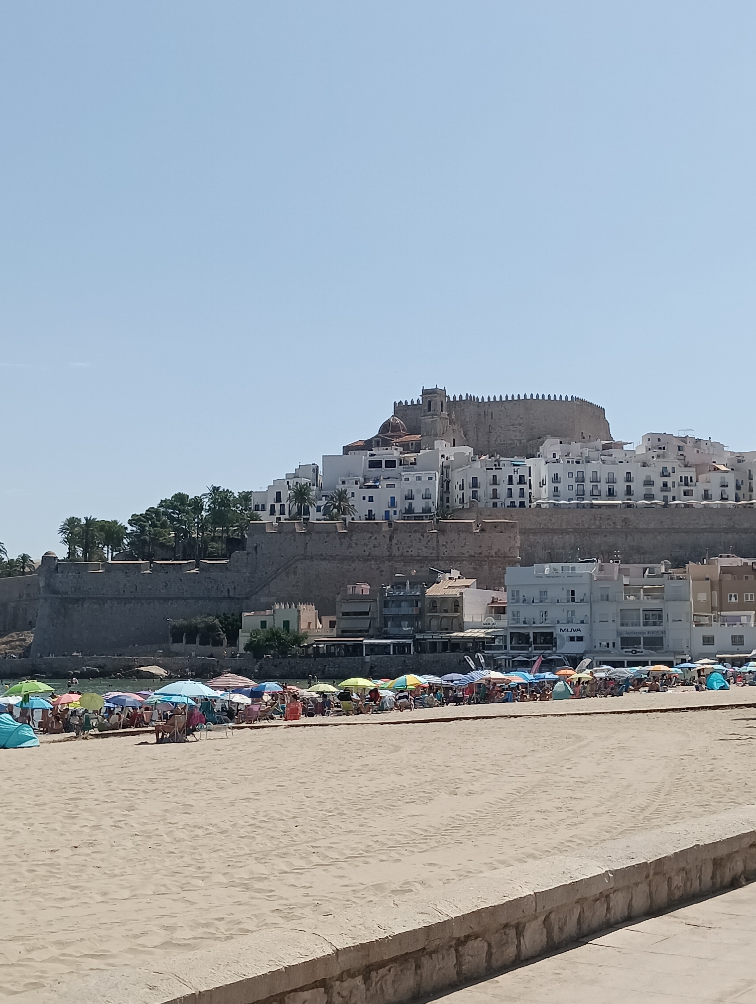 The image captures a sun-drenched scene on the beach in Peníscola, Spain, with the iconic Castillo de Peñíscola perched on a rocky promontory in the background. The foreground is dominated by a wide expanse of sand, marked with tire tracks and lined by a stone wall. Further back, the beach teems with activity. Numerous colorful beach umbrellas and tents are set up, providing shade for beachgoers who are relaxing, sitting, and standing. Some individuals are visible in the water, enjoying the sea. The town of Peníscola rises behind the beach, characterized by its whitewashed buildings with blue accents, characteristic of Mediterranean architecture. The imposing stone walls of the old town and the castle are clearly visible, standing as a testament to the city's historical significance. The sky is a clear, bright blue, indicating fair weather.