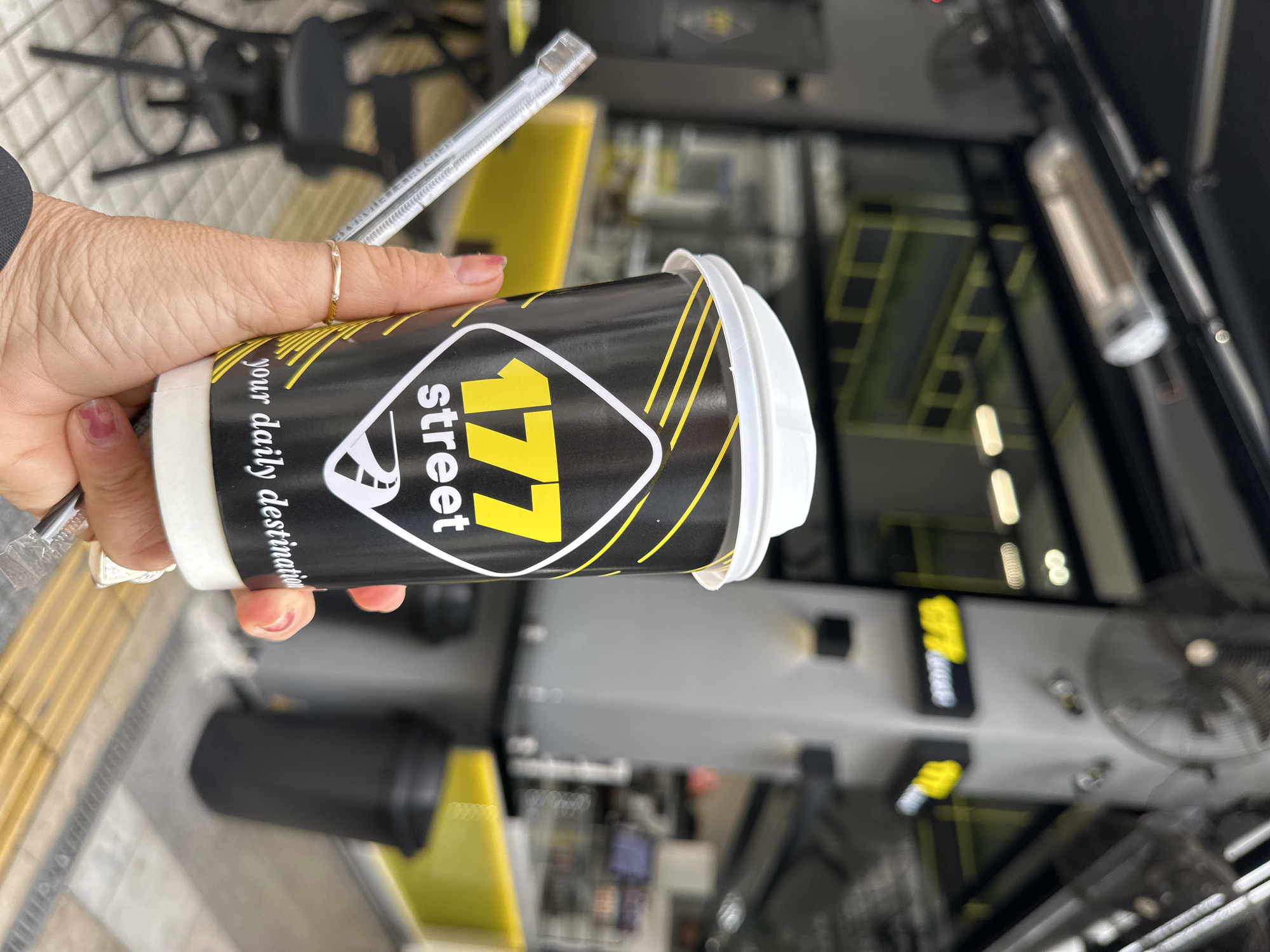 A hand holds a cup with the logo "177 street, your daily destination". The cup is white on top, black in the middle, and features the yellow "177 street" logo in a diamond shape. The hand wears a simple gold ring. A clear stirrer is also in the hand. The background shows an interior scene with a yellow and grey color scheme, reflective surfaces, and elements that suggest an eatery or a cafe, possibly within the city of Serres, Greece, given the brand "177 street" has locations there.