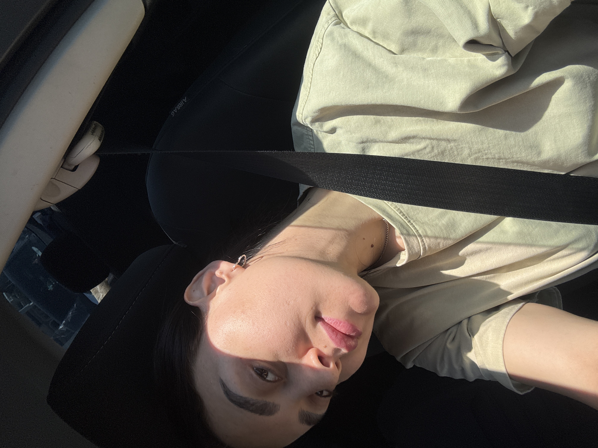 A person with light skin, dark hair, and dark eyebrows, wearing a seatbelt, is sitting in the driver’s seat of a car. The person is wearing a tan-colored t-shirt. The car's interior is visible, including the airbag label on the seat. No information in the image indicates the city is Tbilisi, Georgia.