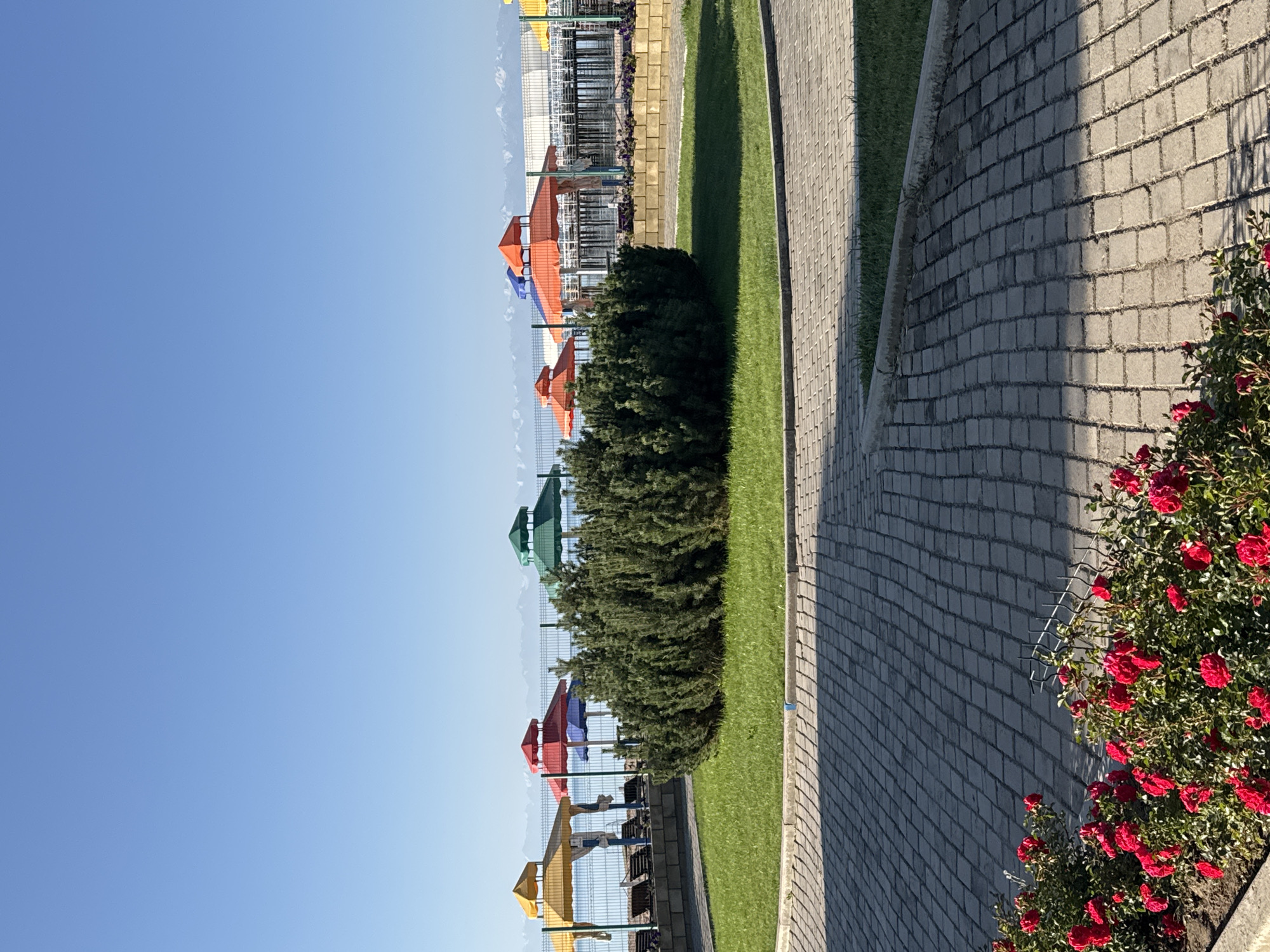 The image depicts a serene outdoor scene likely at a resort in Sary-Oy, Kyrgyzstan, possibly on the shores of Issyk-Kul Lake. In the foreground, a paved walkway made of interlocking grey pavers leads into the scene, bordered by a bed of vibrant red roses. Beyond the walkway is a neatly manicured lawn with a large, dense evergreen bush at its center.
In the middle ground, a fence separates the manicured area from the beach. Behind the fence, a calm body of water, presumably Issyk-Kul Lake, is visible, with distant snow-capped mountains on the horizon under a clear blue sky. Several colorful beach umbrellas – yellow, red, blue, green, and orange – are set up on the beach, suggesting a recreational area. Beach chairs are also visible behind some of the umbrellas.
The overall impression is one of a well-maintained public space designed for relaxation and enjoyment, with the natural beauty of the lake and mountains serving as a backdrop. There are no people or animals visible in the image.