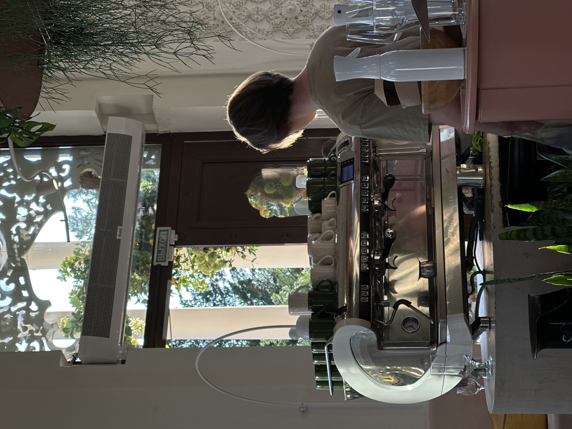 Here is a forensic description of the image:
The image depicts a café setting. A person with short dark hair is operating a large commercial espresso machine, preparing beverages. The machine, which is white and silver, has multiple spouts and a display panel, with several cups hanging from a rack. In the background, a doorway or large window reveals an outdoor scene of trees and foliage. The scene is filled with natural light. A tall, slender white device is affixed to the side of the doorway. There is greenery both in the upper left corner and on the right side of the image, suggesting plants are part of the café decor. The café is located in Unknown, Unknown.