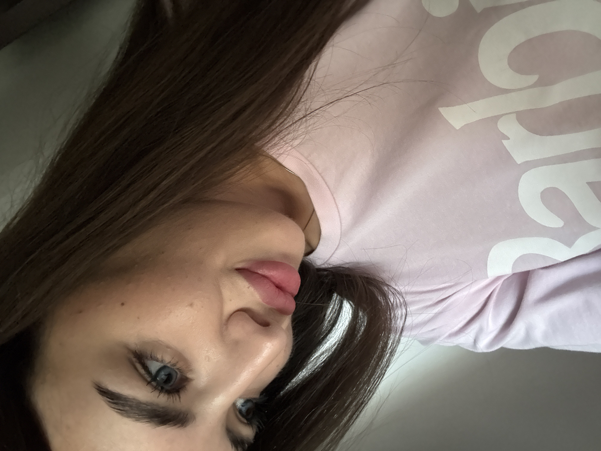 The image shows a close-up, slightly angled portrait of a young woman with long, dark brown hair and blue eyes. She is wearing a light pink t-shirt with the word "Barbie" printed in white. Her head is tilted downwards and to the left, and she is looking slightly off-camera. The background is blurred, appearing to be a neutral indoor setting. There is no visible indication of the city of Chișinău, Moldova.