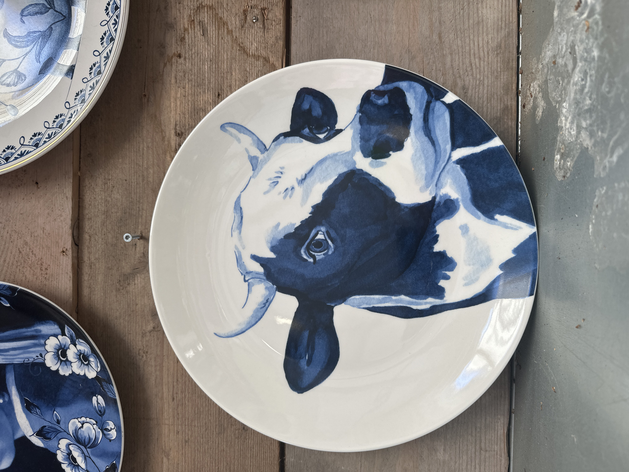 A white ceramic plate with a blue and white painting of a cow's head is displayed against a wooden background. The cow's head is painted in a watercolor style, with varying shades of blue and white. The cow has large, expressive eyes, prominent horns, and a black and white patterned coat. The plate is positioned slightly to the right of the center of the frame. In the background, two other plates are partially visible, one with a floral blue and white pattern and another with a blue and white patterned border. The wooden background appears to be made of planks. The scene is lit from above, casting a slight shadow on the plate. The overall impression is of a decorative display of ceramic art, possibly in a shop or home setting. There is no information to suggest the location is in Tuk, Netherlands.