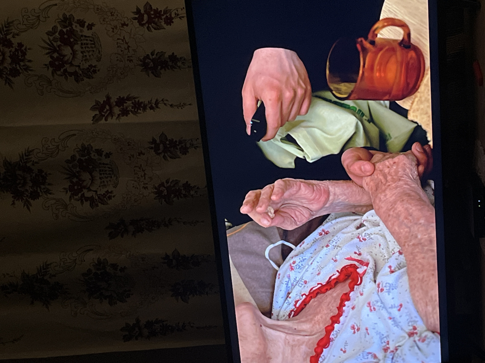The image shows an elderly person in bed, wearing a floral nightgown with red trim. Their hands are visible, one holding a small, light-colored object. Another hand is holding a remote control above a green cloth. Beside the remote control is a small brown pitcher being held by a third hand. The scene appears to be indoors, possibly a bedroom. To the side is a printed cloth. The image appears to be a photo of a television screen. There is no information to indicate the location is Lozarevo, Bulgaria.