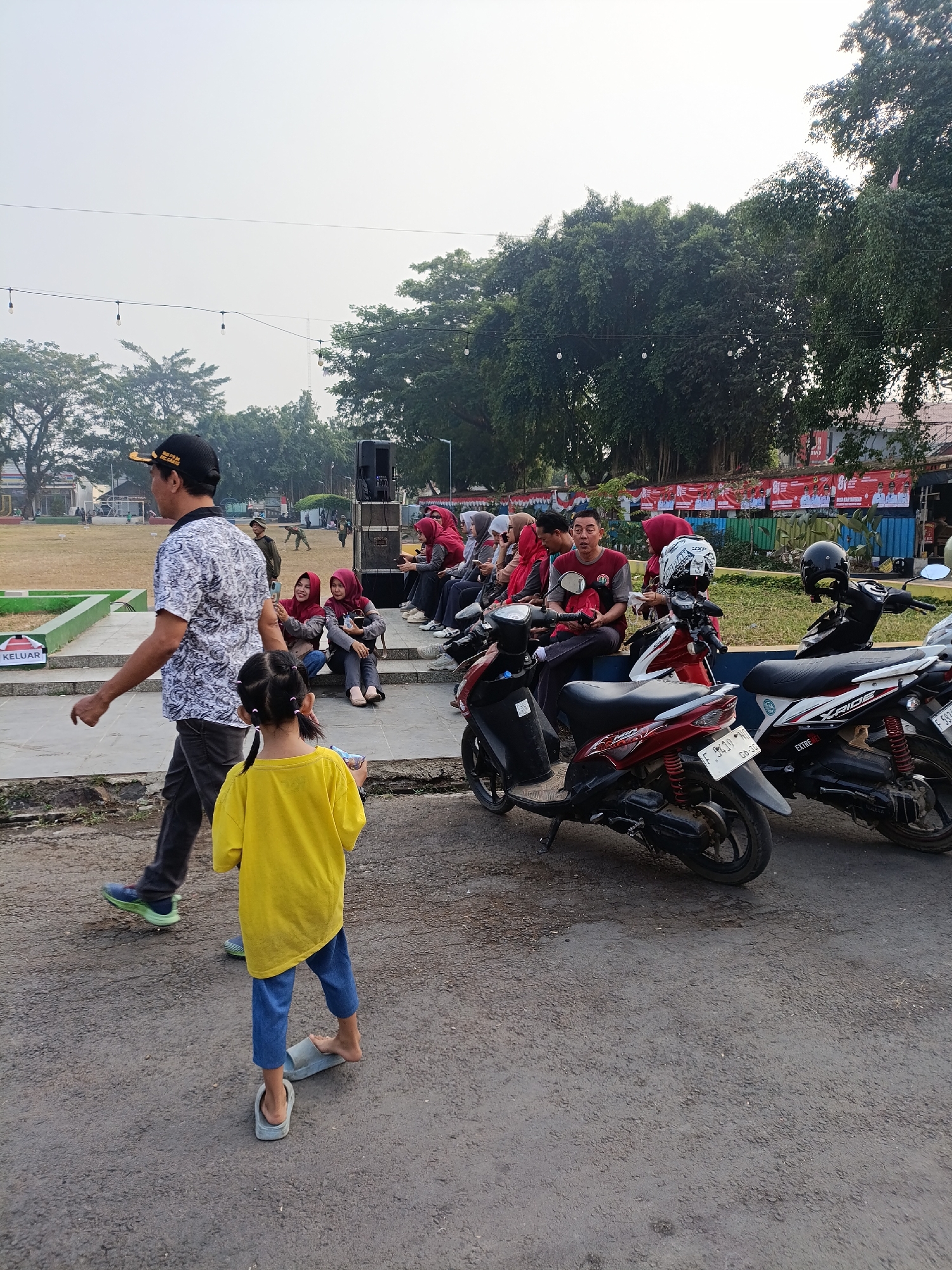 The image depicts an outdoor scene in Cariu, Indonesia, likely during daytime given the bright sky and natural light. The setting appears to be a public area, possibly a park or event space, with a grassy field and trees visible in the background. A group of people are gathered, some seated on a raised platform or steps, while others are standing or walking.
Several motorcycles are parked in the foreground, suggesting this might be an event where attendees arrived by vehicle. The people are dressed casually, with some wearing traditional Indonesian headscarves (hijabs). There's a sense of casual social gathering, with some individuals interacting and others seemingly observing.
A man in a patterned shirt and a cap walks towards the left side of the frame, while a young girl in a yellow shirt and blue pants is in the foreground, facing away from the camera. She is wearing slip-on sandals. In the background, there's a speaker system on a stand, hinting at a possible event involving music or announcements.
Visible text includes a sign on the raised platform that reads "KELUAR" which means "EXIT" in Indonesian. There are also banners with text and images hanging on a wall in the mid-ground, though the text is too small to be legible. The overall atmosphere seems relaxed and communal, with the presence of both adults and a child. The weather appears to be clear.