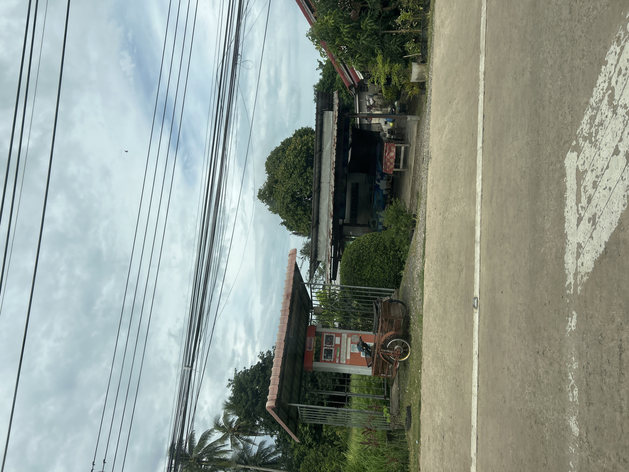 The image, taken in Pa Khlok, Thailand, depicts a roadside scene with a small structure that appears to be a former gas station or vending area. A red and white vending machine, possibly for cigarettes or other small items, is housed under a covered shelter with a corrugated metal roof supported by metal posts and a metal fence. A vintage-looking motorcycle with a sidecar is parked in front of the structure. Adjacent to this, another open-air structure with a similar roof and supports displays tables and what look like goods for sale or storage. The environment is lush and green, with palm trees and other vegetation visible. Overhead, a tangle of electrical wires crisscrosses the sky, which is partly cloudy. The road in the foreground is asphalt with a white line marking. No people are visible in the image.