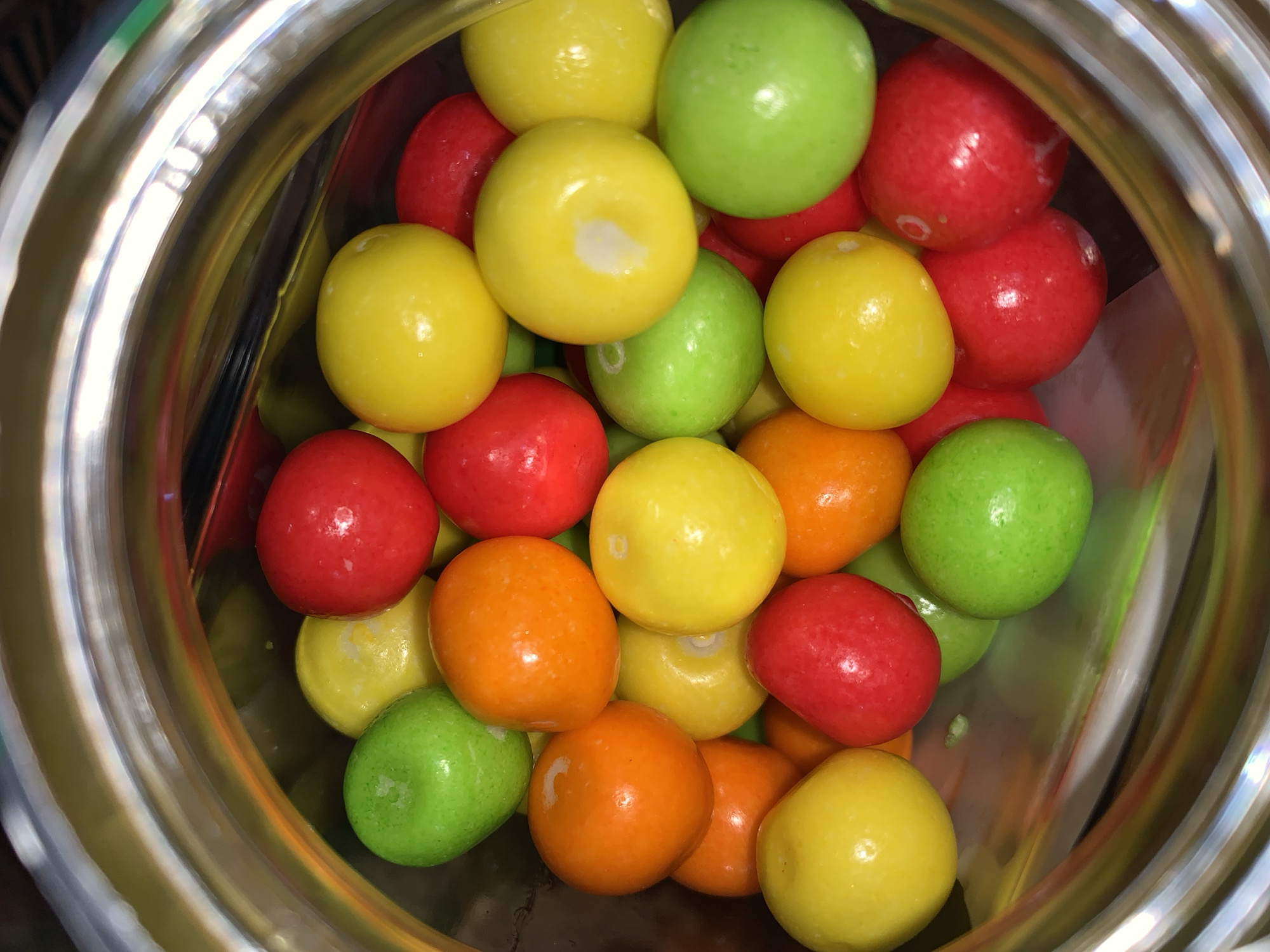 A high-angle, close-up view captures a dense collection of brightly colored, spherical candies inside a clear, cylindrical container. The candies are primarily red, yellow, green, and orange, exhibiting a glossy, uniform finish. They are tightly packed, filling the majority of the frame, with the clear, reflective rim of the container visible in the upper and lower portions, creating concentric bands of light and subtle reflections. No individuals are present. The environment consists solely of the container and its confectionery contents.