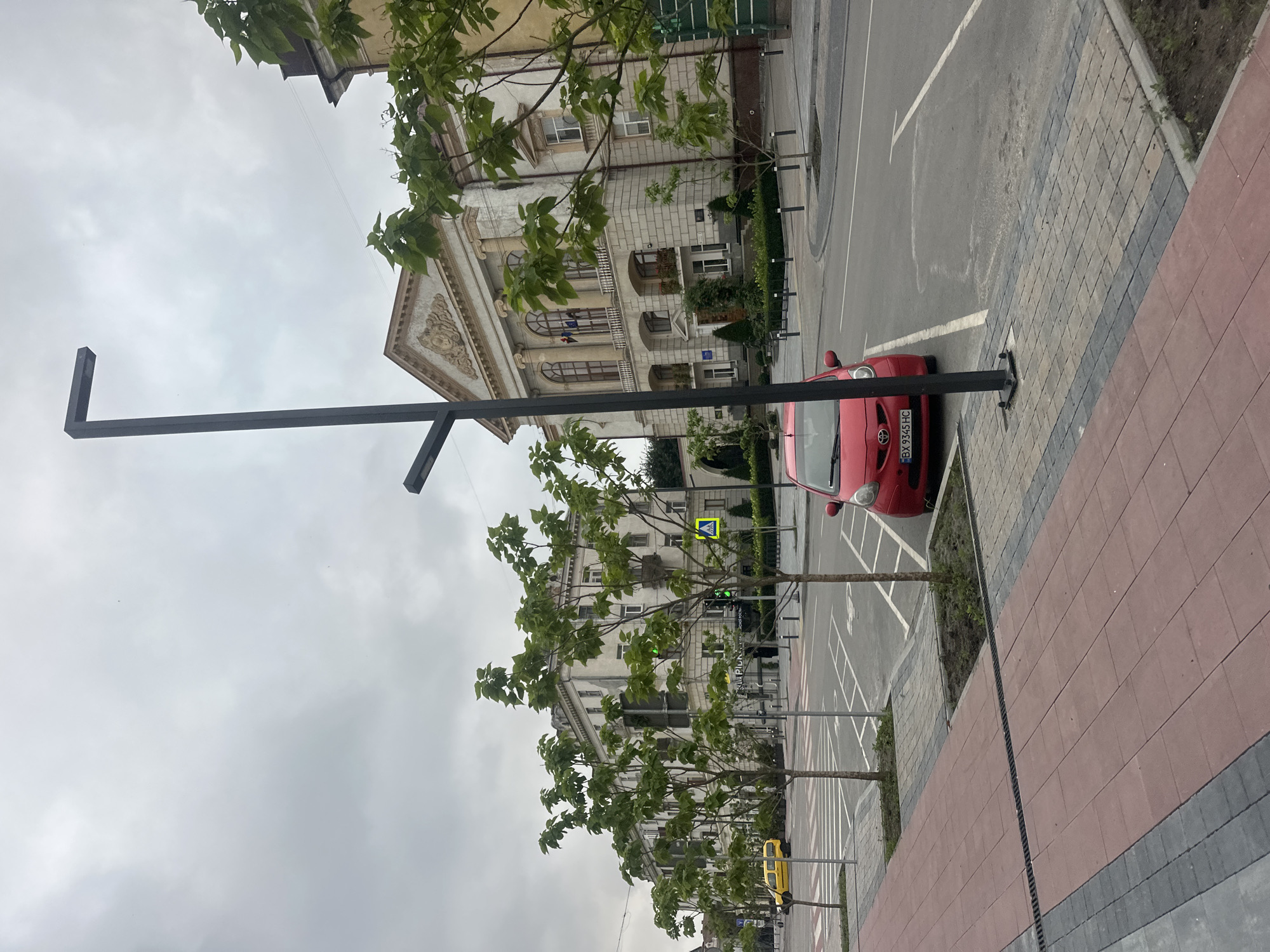 The image shows a street scene in Ternopil, Ukraine, under an overcast sky. In the foreground, a red hatchback car is parked in a designated parking space. The car has a Ukrainian license plate reading "BX 9345 HC". Adjacent to the parking area is a pedestrian walkway with red and grey paving stones.
Further down the street, a row of young trees with green foliage lines the sidewalk, separating the pedestrian area from the road. Behind the trees, a long, multi-story building with a light-colored facade stands prominently. The building features classical architectural elements, including arched windows, columns, and ornate decorative details above the main entrance. Flags are visible on the upper floors of the building.
Across the street from the main building, another row of similar buildings can be seen. In the distance, a yellow van is visible, and further down the road, a pedestrian crossing with a green traffic light is present. Modern streetlights with a dark finish are also visible throughout the scene, indicating a well-maintained urban environment. The overall impression is a quiet street in a city, likely during daylight hours given the illumination.