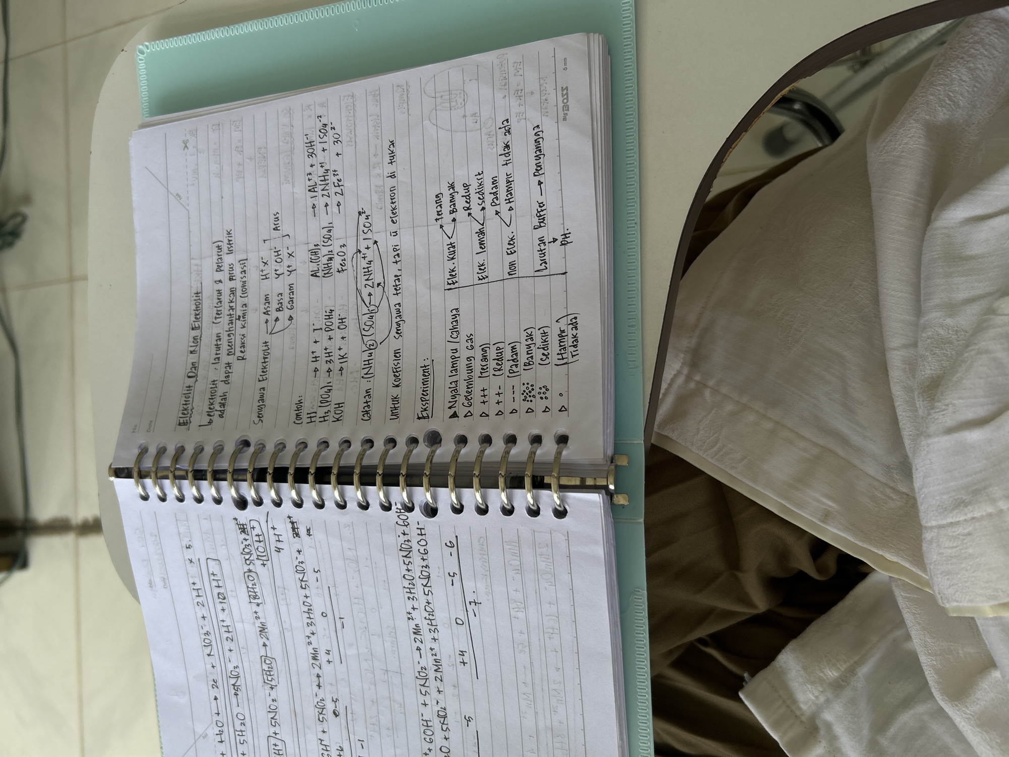 An open, spiral-bound notebook, rotated 90 degrees clockwise, rests on a light-colored surface, likely a desk. The notebook has a light blue plastic binder cover on the left. The pages are filled with handwritten notes, diagrams, and chemical equations, primarily in black ink. Part of a white and brown fabric, possibly clothing or bedding, is visible in the blurred background.
The visible notes are in Indonesian and pertain to chemistry, specifically "Elektrolit Dan Non Elektrolit" (Electrolytes and Non-Electrolytes). Definitions explain that electrolytes are solutions that can conduct electric current through chemical reactions (ionization). Examples of acids, bases, and salts are given with their ionization reactions, such as "AL(OH)3 → AL+3 + 3OH-" and "(NH4)2(SO4) → 2NH4+ + SO4-2". A note mentions that compound coefficients remain constant, but electrons are exchanged.
An "Eksperimen" (Experiment) section details observations related to the strength of electrolytes, including "Nyala lampu/cahaya" (light bulb/light) and "Gelembung Gas" (gas bubbles), with categories like "Flek. Kuat" (strong spot), "Flek. Lemah" (weak spot), and "non Elek." (non-electrolyte), described by terms such as "terang" (bright), "redup" (dim), "padam" (off), "banyak" (many), "sedikit" (few), and "Hampir tidak ada" (almost none). The term "Larutan Buffer → Penyangga" (Buffer Solution → Buffer) and "pH" are also visible. The left page contains complex chemical equations, likely redox reactions, with coefficients and oxidation states indicated (e.g., "+4", "0", "-5", "-1"). The brand name "DIS BOSS" and "6 mm" are visible on the bottom right page.