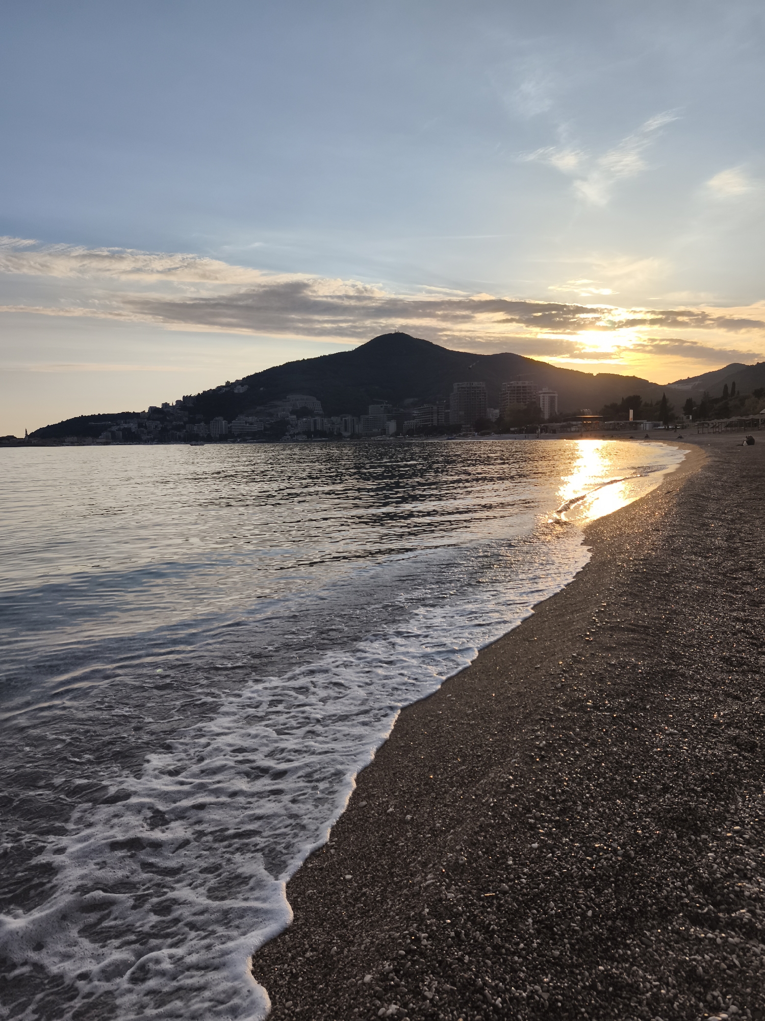The image presents a tranquil sunset scene over the coast of Budva, Montenegro. The sun, a bright golden orb, is low on the horizon, nestled behind distant mountains on the right, casting a warm, radiant glow across the landscape. The sky above is a blend of soft blue, transitioning into delicate hues of orange and yellow near the horizon, with wispy, scattered clouds.
In the foreground, the gentle waves of the sea break upon a dark, pebbled beach. The frothy white foam of the waves creates a dynamic edge where the water meets the shore, and the wet pebbles brilliantly reflect the golden light of the setting sun. The sea itself appears calm, with ripples and shimmering reflections stretching towards the horizon.
Across the midground, a series of mountains rise, silhouetted against the bright sky. Along their base and slopes, numerous buildings, likely hotels or apartments, dot the coastline, indicating a developed coastal area. These structures add a subtle human element to the otherwise natural vista. The perspective is low, drawing the viewer into the scene along the curving coastline.
The time of day is clearly sunset, creating a peaceful and serene atmosphere. There is no discernible specific activity taking place, and no visible text can be identified in the image.