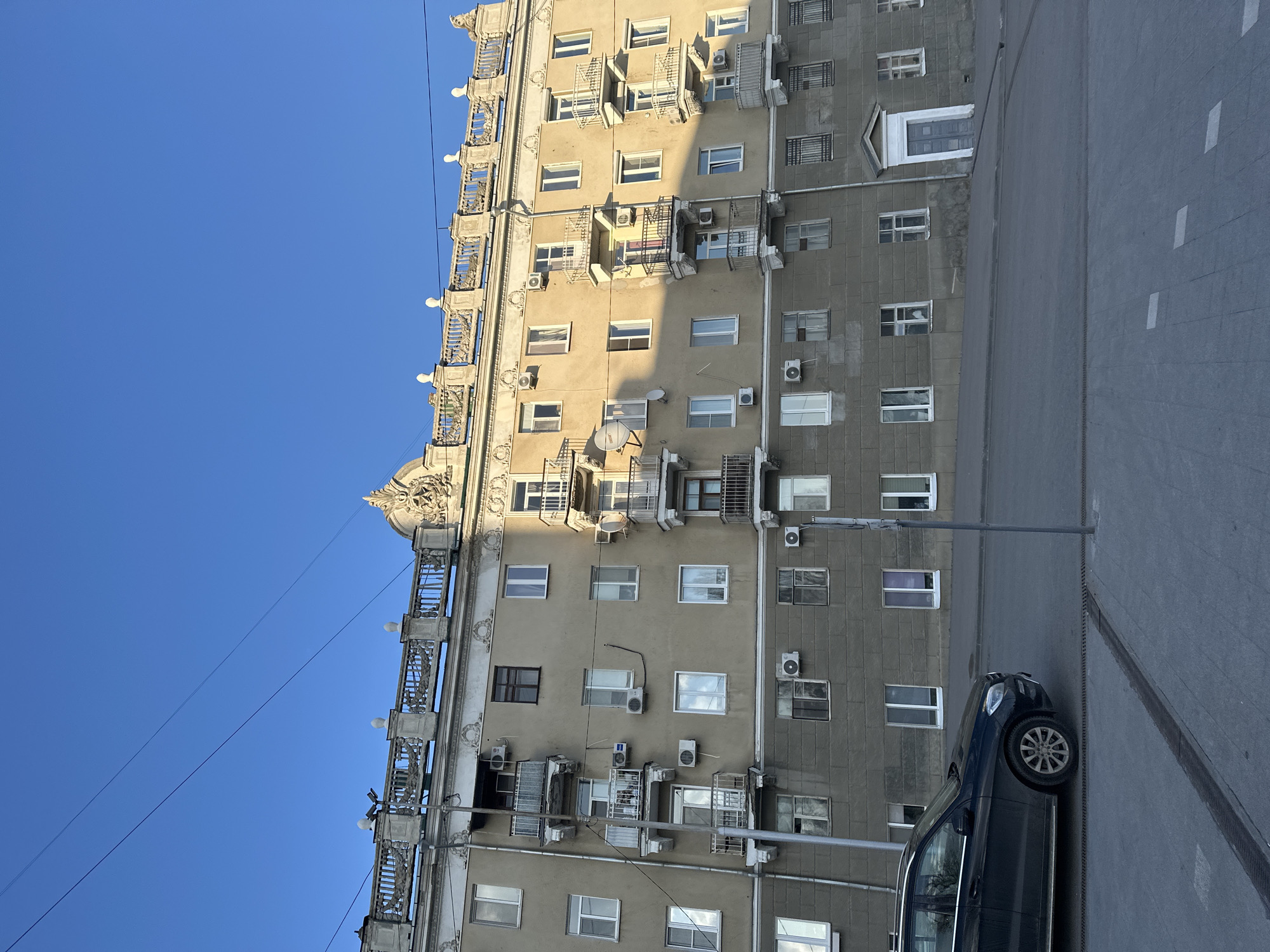 The image presents a detailed view of a multi-story urban building under a clear blue sky, likely in Saratov, Russia. The building occupies the majority of the frame, showcasing two distinct facade sections: a light beige or tan section on the left, and a darker, textured gray section to the right.
The beige facade is architecturally ornate, featuring decorative balconies or window structures adorned with intricate railings and lamps, particularly noticeable along its left edge. A sculpted, embellished element crowns the top left corner of this section. Both facades are punctuated by numerous windows, many with white frames, suggesting residential units. A prominent feature across the entire building are the numerous external air conditioning units and several satellite dishes mounted on the walls. Some windows on the gray facade are also fitted with security bars or grills.
The lighting indicates direct sunlight, suggesting a late morning or early afternoon time, with shadows visible on the building's surface. The sky is a vivid, cloudless blue, with thin utility wires stretching across it. A slender street light or utility pole stands vertically between the building and the paved area below.
In the lower right portion of the image, a dark-colored SUV or sedan is parked on a paved surface, which appears to be a street or a wide sidewalk. White painted lines are visible on this surface, along with a storm drain or grate system. No specific activity or individuals are depicted, offering a static glimpse of an urban environment. There is no visible text in the image.