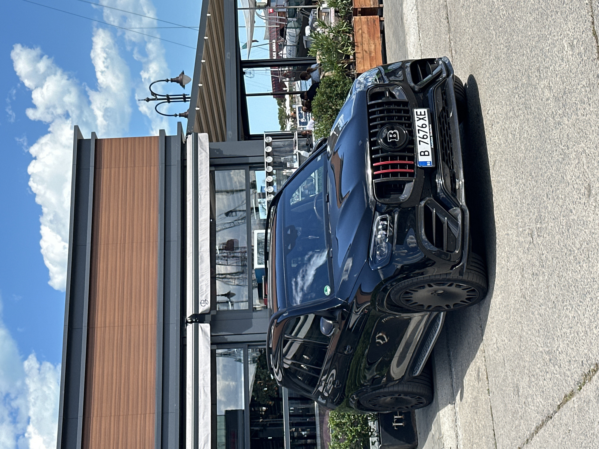 The image shows a black Brabus SUV parked on a sidewalk in Varna, Bulgaria, evident from the Bulgarian license plate "B 7676 XE". The SUV is parked in front of a modern building with a wood-paneled section and a glass-walled restaurant area. People are visible inside the restaurant. The sky is blue with scattered clouds.