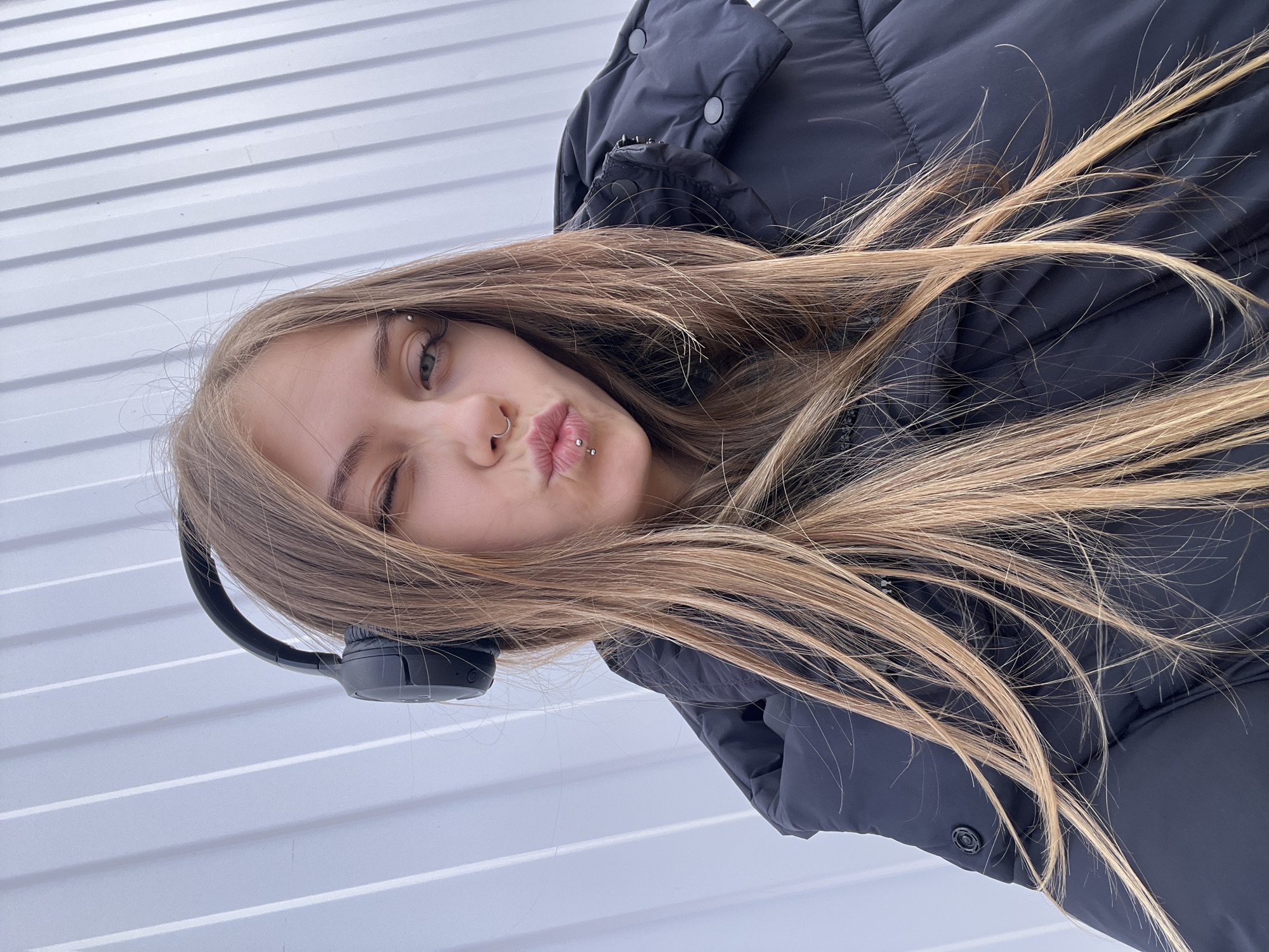 A young female is the central subject of this close-up portrait, captured from a slightly high angle, likely a selfie. She has long, straight hair with shades of light brown and blonde, which flows over her shoulders and part of her black puffer jacket. She wears black over-ear headphones, with the left ear cup visible near her ear. Her face features several silver-toned piercings: one in her right eyebrow, a septum ring, and a labret piercing below her lower lip. She is winking with her left eye and pursing her lips in a playful, almost "kissing" expression. The background consists of a plain, light-colored, possibly white or light grey, corrugated metal wall with distinct horizontal lines, providing a simple and uniform backdrop. The lighting is bright and even, suggesting daytime conditions, possibly overcast, which aligns with the warm clothing worn. The overall mood conveyed by her expression is confident and lighthearted. There is no visible text within the image. The location context provided for this image is Фоки, Russia.