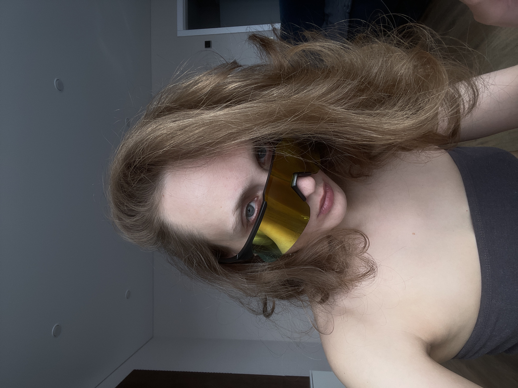 A close-up, tilted portrait features a young individual with light brown, wavy hair. The subject's face is largely obscured by large, wrap-around sunglasses with reflective gold-yellow lenses and a black frame. The visible features include a blue-gray left eye, the bridge of the nose, and light pink lips. The person wears a dark, possibly gray or black, strapless top.
The setting is indoors, characterized by plain, light-colored walls, which appear gray under the current lighting. Several circular fixtures, resembling recessed lights or smoke detectors, are visible on the walls. A portion of a white window frame can be seen in the upper right. A dark, indistinct object, possibly furniture or a rug, occupies the bottom right corner.
The activity is a posed photograph, likely a selfie, given the close proximity to the subject and the direct gaze of the visible eye. The subject's expression is neutral. The lighting suggests an indoor setting, possibly with ambient or artificial light, casting soft shadows. There is no visible text in the image. The location context for this media file is Podshivalovo, Russia.