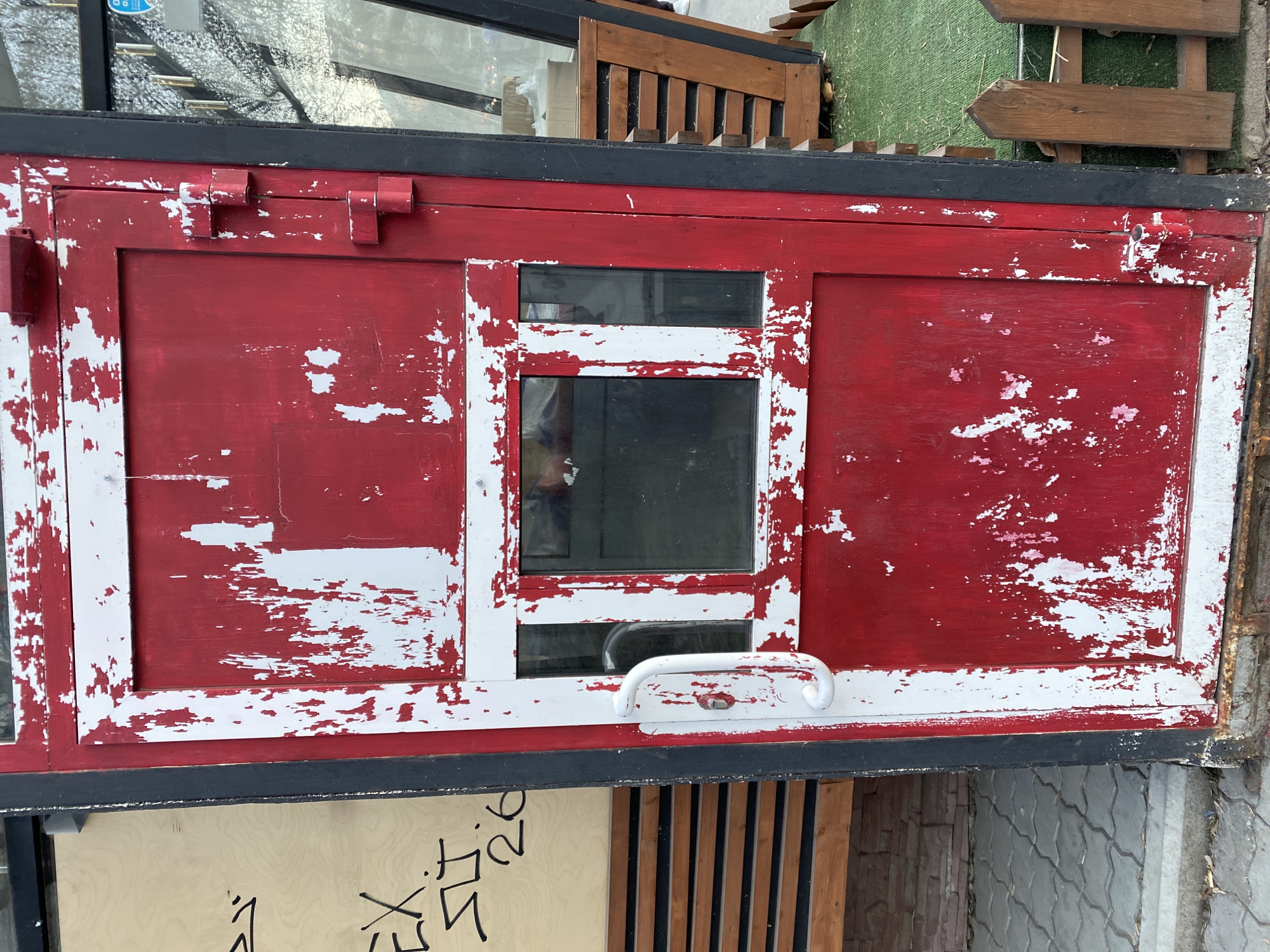 This image captures a weathered, red and white structure, likely an outdoor cabinet, kiosk, or small storage unit. The dominant color is red, but significant portions of white paint are visible underneath, exposed by extensive chipping and peeling, creating a distressed aesthetic. Black trim frames the top and bottom edges of the structure.
In the center of the red surface, two rectangular glass panes are set into a white frame. Reflections in the glass show indistinct background elements and what appears to be a person taking the photograph. Below the lower glass pane, a white, curved handle is prominently featured. On the top left of the red surface, there's a small, red, box-like attachment, and on the top right, two red hinges suggest a door opening to the right.
The setting appears to be outdoors during daytime, with bright, even lighting. Above the red structure, elements like a wooden slatted fence or bench back, green artificial turf, and a wooden directional arrow sign are visible, hinting at a public or recreational area. Below the structure, a light-colored wooden panel displays black handwritten text, where the numbers "26" are clearly discernible amidst other less legible symbols or characters. Further below, patterned gray and brown paving stones are visible. The location, as per context, is Yekaterinburg, Russia. No specific activity or emotion is depicted.
