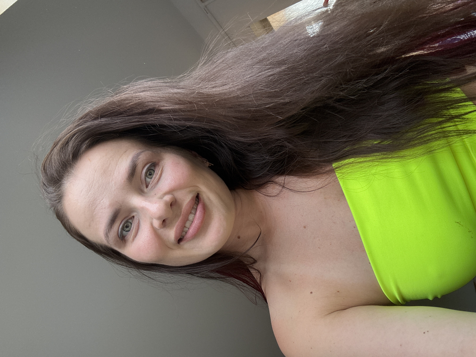 A woman with long, dark brown hair and fair skin is shown from the chest up, looking directly at the camera. She is wearing a bright lime green strapless top. Her eyes are green and she has a slight smile. The background appears to be an indoor setting with a plain, gray wall. There are no discernible landmarks or city-specific features.