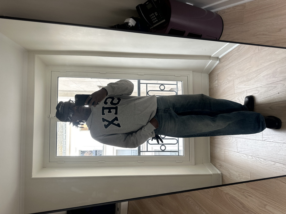 A person is captured in a full-length mirror selfie, standing indoors and facing towards the camera. The individual is wearing a light gray hooded sweatshirt with the word "SEX" printed in large, black, uppercase letters, appearing reversed due to the mirror reflection. They have one hand in the pocket of their baggy, light-wash blue jeans. Black shoes are visible on their feet. On their head, they wear a dark cap and glasses, while holding a dark-colored smartphone up to take the photo.
The indoor environment features light-colored wooden floorboards and plain light-colored walls. A large window or French door occupies the background, revealing an outdoor scene. The window has a white frame with multiple panes, and a classic wrought iron balcony railing is visible directly outside. Beyond the railing, typical Parisian Haussmannian buildings with light-colored stone facades, multiple windows, and sloping gray roofs are visible under what appears to be daylight conditions. A purple, multi-tiered bin with electronic devices, including what resembles a landline phone, is partially reflected in the mirror on the right side.
