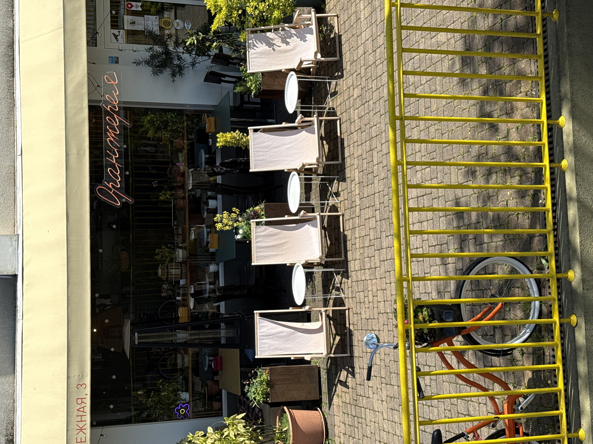 The image captures the exterior of a cafe or establishment from an elevated perspective, rotated 90 degrees clockwise. The scene is bathed in bright daylight, casting strong shadows, indicating clear weather.
The left side of the image features the cafe's glass-fronted facade, protected by a cream-colored awning. Inside, potted plants and reflections of the street are visible. A prominent orange neon sign in a cursive script spells out "Гранитерра" (Graniterra), likely the name of the business. To the far left, a vertical sign on the building reads "ЕЖНАЯ, 3" (EZHNAYA, 3), suggesting a street name and building number. Various smaller logos and stickers, including what appears to be a "24/7" sign and a Wi-Fi symbol, are present on the entrance door. A blue/purple neon flower sign illuminates from within the cafe.
In front of the cafe, several outdoor seating arrangements are set on a patterned brick-like pavement. These include four sets of light-colored, fabric-seated chairs with wooden frames, resembling deck chairs, each paired with a small round white table on a metal stand. Additional potted plants are placed along the cafe's exterior.
On the right side of the image, a bright yellow metal fence or barrier runs parallel to the cafe. It consists of multiple horizontal bars and is anchored by yellow caps at its base. An orange bicycle, with a silver handlebar and white-rimmed wheels, is parked behind this fence, partially obscured. No people are visible in the scene, suggesting a quiet moment for the establishment.