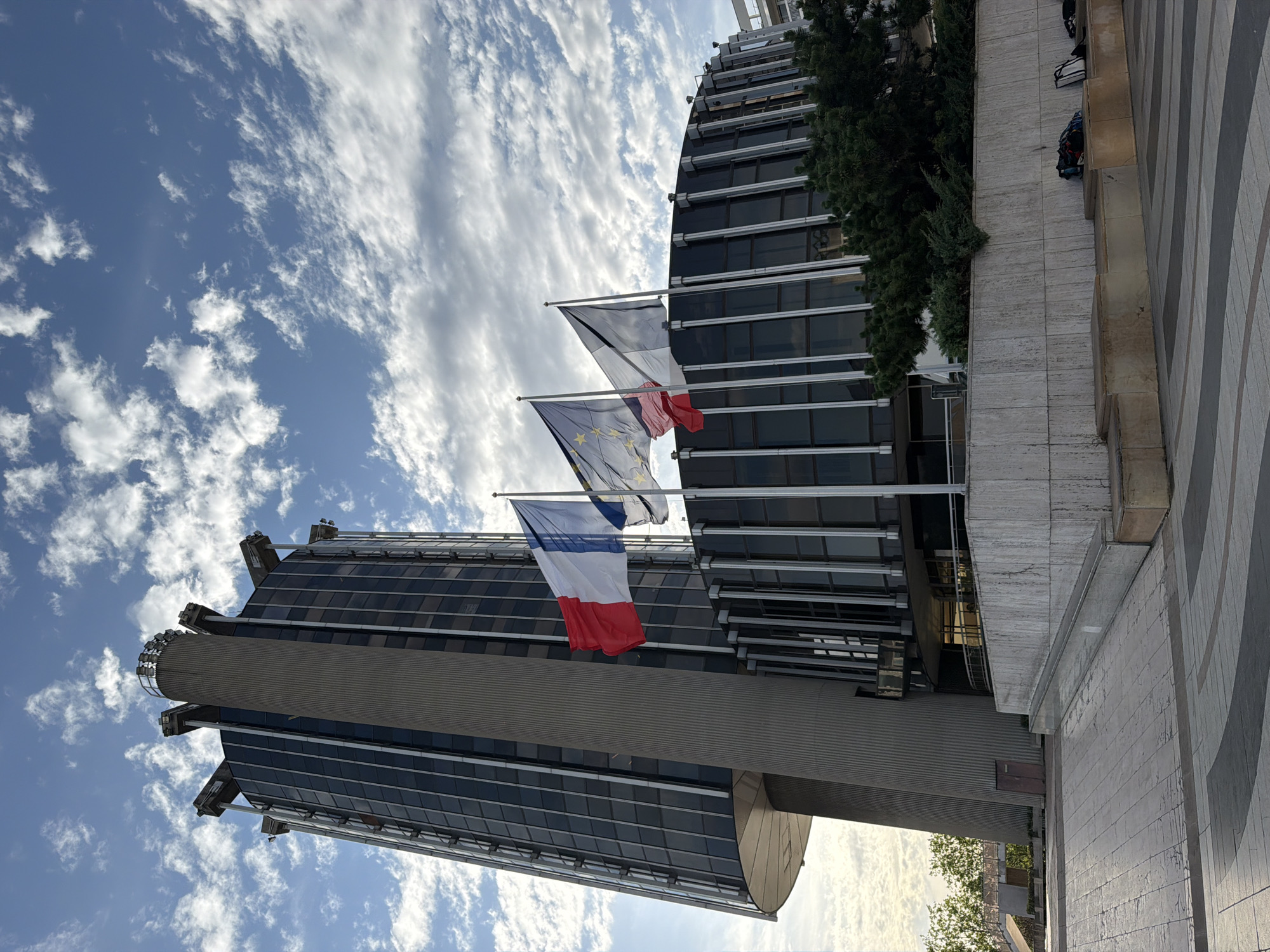 A medium-shot, slightly low-angle, exterior, daytime photograph shows the Palais de Justice in Créteil, France. The building is a tall, cylindrical tower with a concrete core and glass-paneled exterior, topped with a circular structure featuring multiple spotlights. To the right of the tower is a curved, multi-story section of the building, also with dark glass panels. In front of the curved section, three flags are flying: the French flag on the left, followed by the flag of the European Union, and then another flag, possibly a regional flag, on the right. The flags are blowing in the wind. The building is situated in a paved plaza with a patterned ground surface. A low, stone retaining wall with built-in seating runs along the foreground, with several bags placed on the benches. Lush green bushes and trees are visible behind the wall. The sky above is a bright blue with scattered white clouds.