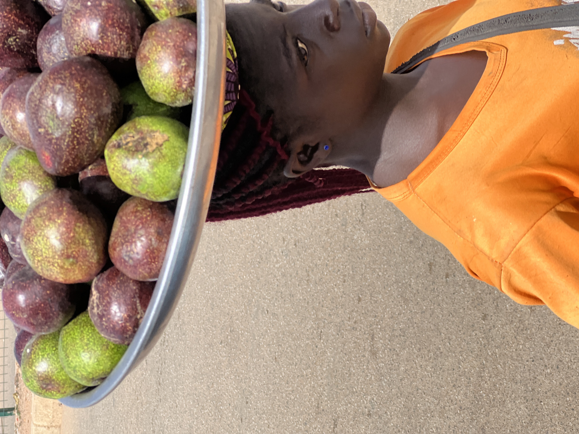 A medium-shot, eye-level image shows a woman standing on a paved surface. The woman has brown skin and wears an orange t-shirt. She has maroon braids, a blue stud earring in her left ear, and a colorful head covering. She carries a large silver bowl filled with what appears to be a variety of round fruits in shades of green and maroon. The setting is likely Bukuru, Nigeria.