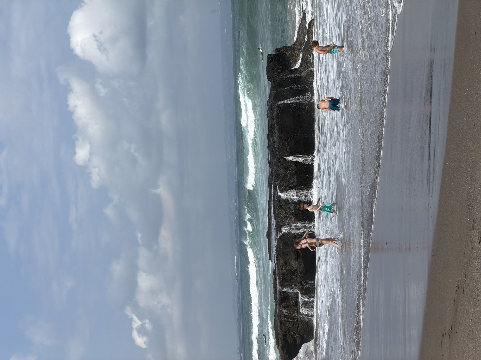 This media file captures a scene at North Kuta, Indonesia, with a focus on a rocky coastline during what appears to be a sunny day, judging by the bright sky and scattered clouds. The sea is active with moderate waves crashing against dark, wet rocks.
The primary subjects are several individuals interacting with the coastal environment. There are at least four people visible, most of whom are wearing swimwear. One person, a male with light hair, is standing on the rocks, wearing light blue patterned swim trunks. Another individual, also male, is lying on their stomach on the rocks, wearing dark blue swim trunks. A third person, appearing to be female with dark hair, is standing in the shallow water near the rocks, wearing a light-colored swimsuit. A fourth person, a male with light hair, is seen standing further back on the rocks, wearing light blue swim trunks.
The activity taking place is people enjoying the beach and ocean. They seem to be exploring the rock formations, wading in the water, or simply relaxing by the sea. The presence of waves suggests conditions suitable for swimming or simply experiencing the ocean's power.
Notable details include the texture and color of the rocks, which are dark and appear to be covered in seaweed or algae in places. The water is a rich green, transitioning to white foam where the waves break. The sky is a mix of blue and white clouds, with some areas appearing brighter than others, hinting at intermittent sunshine. The overall mood seems relaxed and recreational. There is no visible text in the image.