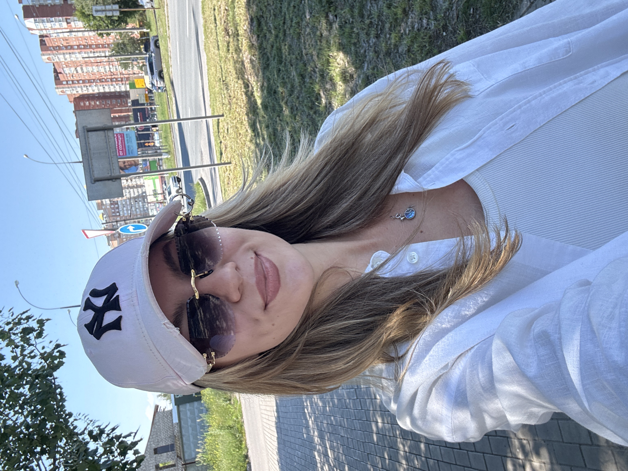 A blonde woman is taking a selfie outdoors in an urban environment. She is wearing a white button-down shirt over a white tank top, a light pink baseball cap with a black NY logo, and rimless sunglasses with gold accents. A small, circular pendant with a blue stone hangs from a silver chain around her neck. Her hair is long and light brown. In the background, there is a paved walkway, a grassy area, a street with cars, and multi-story brick buildings. The sky is blue with scattered clouds.
