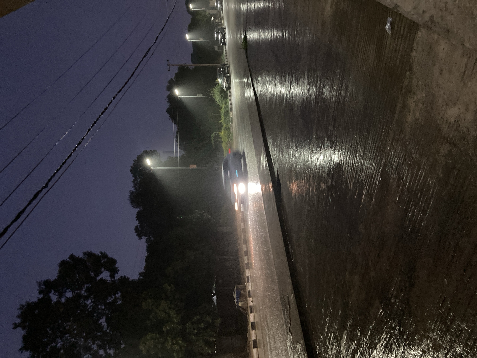 A street scene at night in Jos, Nigeria, during a heavy rain. The road is wet and reflects the light from streetlights and car headlights. Several cars are visible, driving through the rain. Trees line the side of the road, their silhouettes dark against the stormy sky. Overhead, power lines stretch across the frame. The overall atmosphere is dark and moody, with the rain creating a sense of atmospheric visibility.