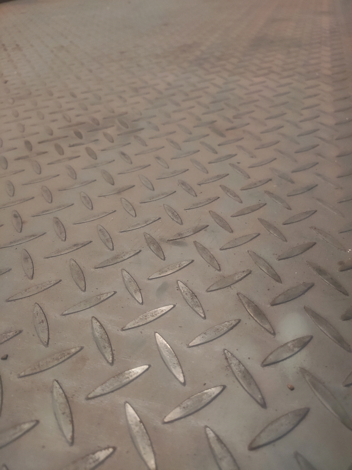 An overhead, close-up, eye-level shot shows a textured, diamond-plate metal surface. The metal is a dull, gunmetal gray with some light streaks and smudges indicating wear and tear. The diamond pattern consists of raised, elongated ovals that create a grid across the surface. The foreground is in sharp focus, with the texture of the metal clearly visible, while the background fades into a soft blur, suggesting a shallow depth of field. There are no people or living subjects visible in the image. The scene appears to be an interior or outdoor industrial or utilitarian setting, such as a factory floor, a workshop, or a loading dock. The lighting is even and diffuse, with no strong shadows.