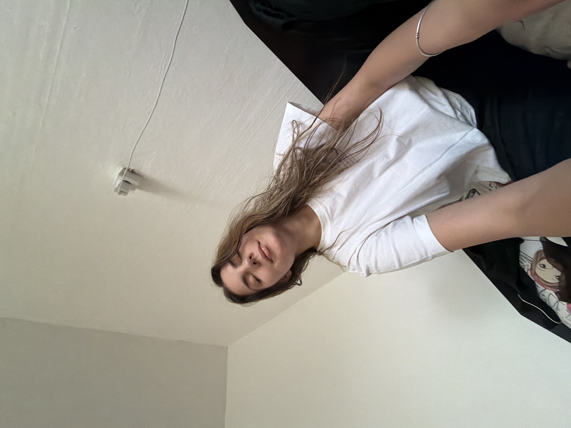A person is lying on a bed, seemingly taking a selfie. The background shows a white wall and a ceiling light fixture. The location is not discernible from the image.