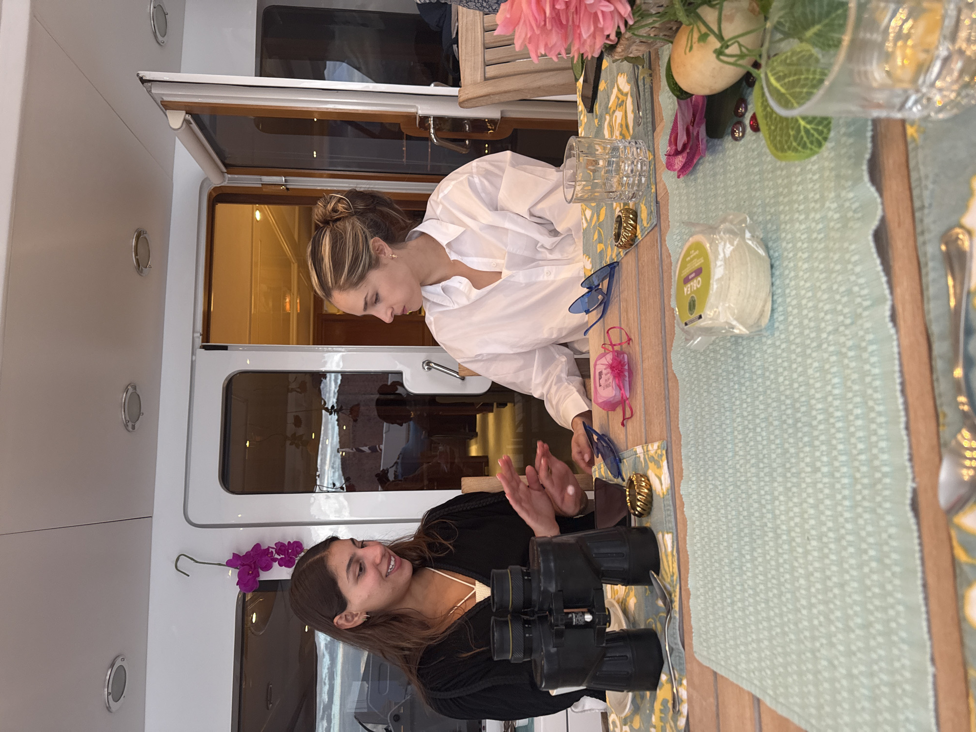 Two women are seated at a table. One woman is wearing a white button-down shirt, and her hair is pulled back in a bun. The other woman is wearing a black cardigan and a white top. On the table are binoculars, a spool of twine, a set of glasses, blue sunglasses, and pink flowers, along with what appears to be a table setting. The scene appears to be on a boat, as evidenced by the windows and wall fixtures. The location is Unknown, Mexico. The women are engaged in a discussion, with one woman clapping.
