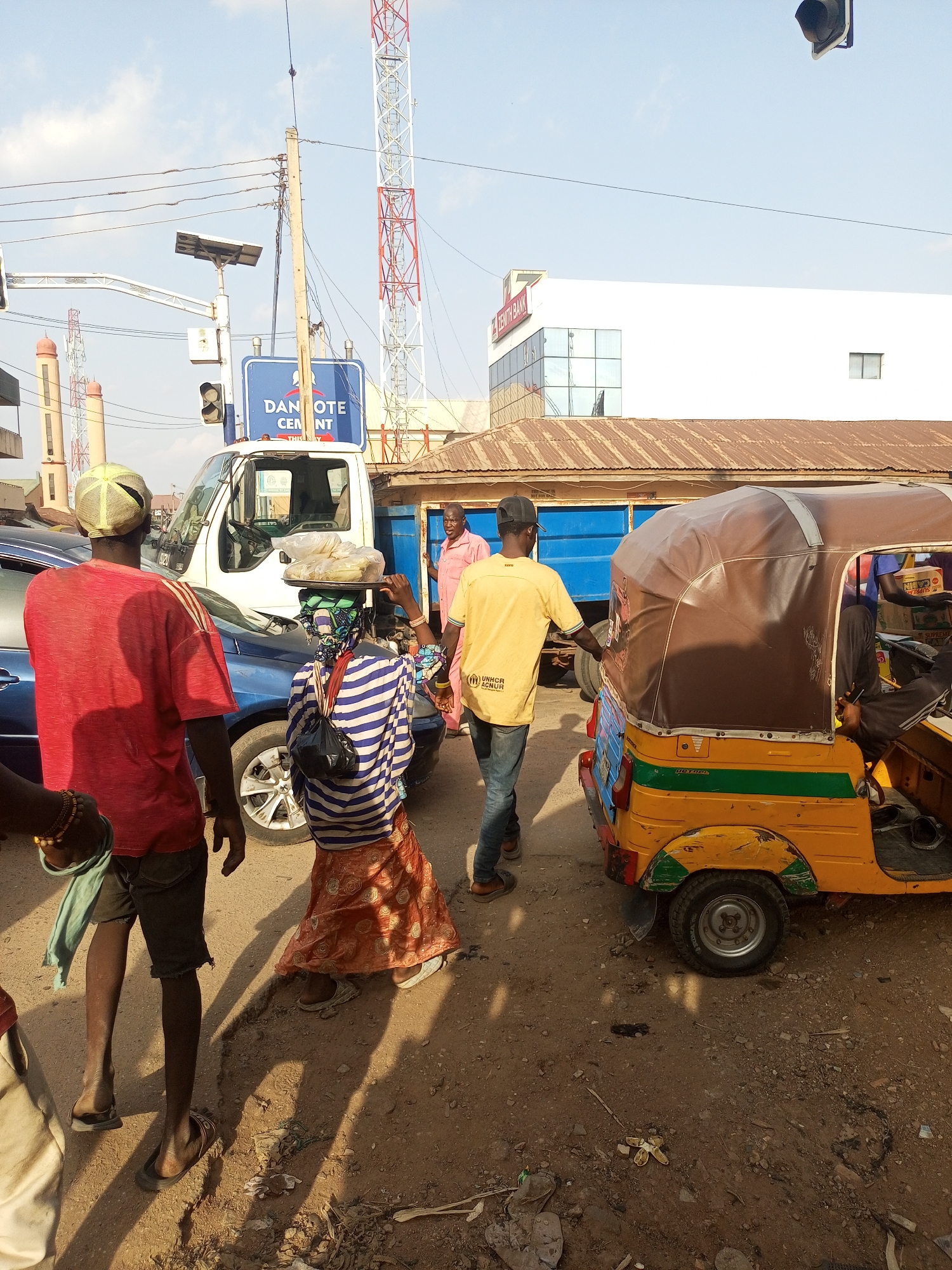 The image depicts a busy street scene in Maiburiji, Nigeria. Several people are walking near a three-wheeled vehicle, known locally as a keke. A woman carries goods on her head. A "DAN GOTE CEMENT" sign is visible. A white truck, blue sedan, and Zenith Bank building are also in the background. A man wearing a yellow shirt is seen moving towards the keke. The ground is dusty. A tall red and white communication tower is visible.