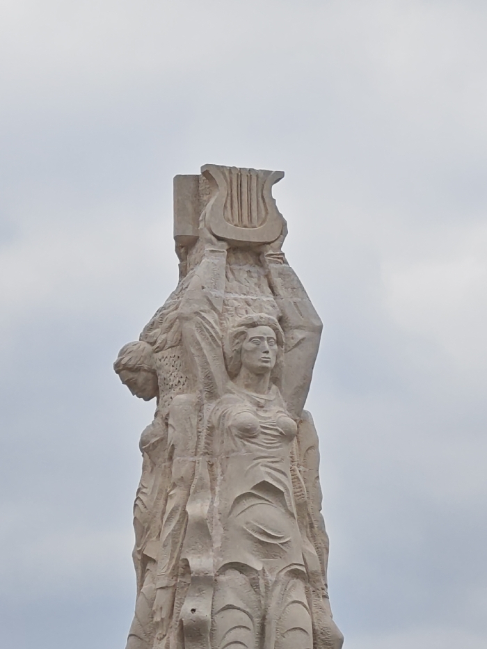 A monumental, light-colored stone sculpture, a detail of the "Monument to 1300 Years of Bulgaria," is depicted against an overcast, light grey sky in Shumen, Bulgaria. The central foreground features a female figure, carved from the same monolithic block, facing slightly right with a neutral expression. She wears a draped, textured garment with prominent breast detail and intricate lower body folds. To her left and slightly behind, another figure's head is visible in profile, facing left and downwards, with textured hair or headwear. Above these figures, a stylized lyre is held aloft by integrated arms, forming the uppermost visible section of the monument. The sculpture exhibits a rough, chiseled texture throughout. No other objects or individuals are present in the immediate visible environment besides the monument and the sky.