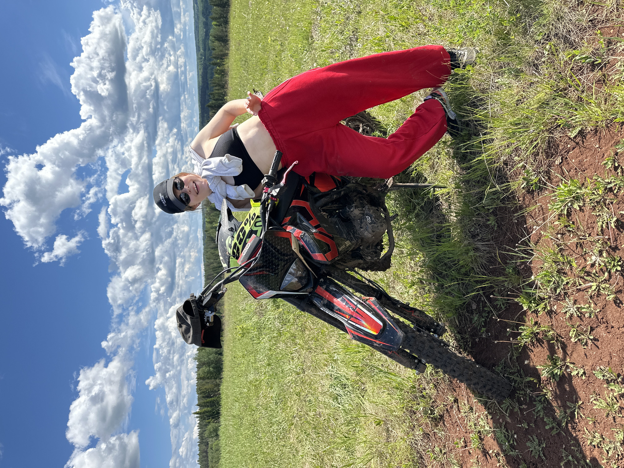 A person is posing on a dirt bike in a grassy field under a partly cloudy sky. The person is wearing a black sports bra, red sweatpants, a white shirt tied around their neck, a black cap, and sunglasses. They are smiling and holding up two fingers. The dirt bike is black and red with mud splattered on it. The field is green with some brown patches of dirt. In the background, there is a line of trees under the blue sky with white clouds. The image seems to have been taken near Пепеляева, Russia due to the presence of trees and grass.