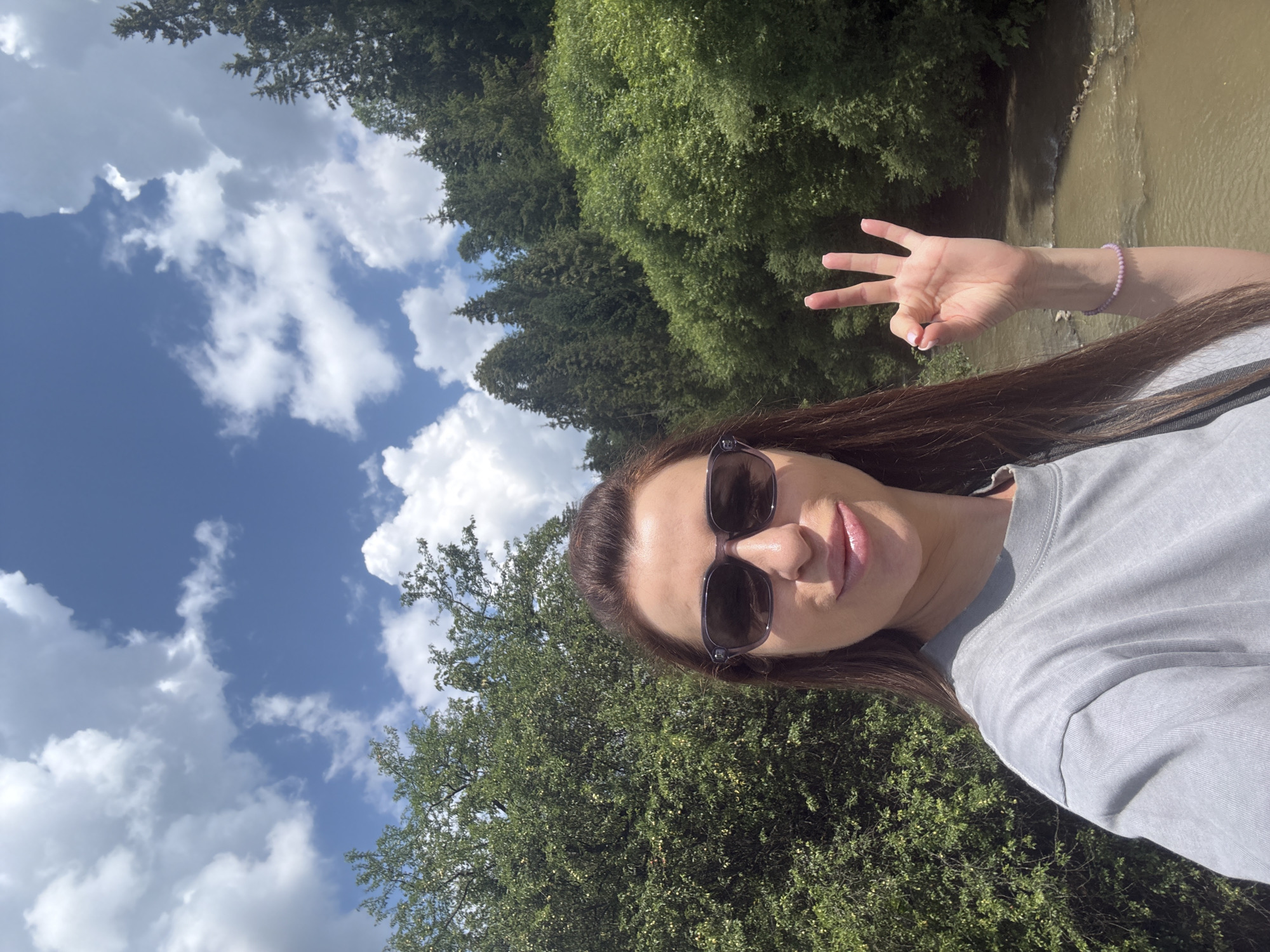In Slavske, Ukraine, a woman with long brown hair and sunglasses takes a selfie outdoors. She is wearing a grey t-shirt and a light purple beaded bracelet on her left wrist. She is holding up her left hand in a gesture with three fingers extended. Behind her, lush green trees and a body of water are visible under a bright blue sky with fluffy white clouds. The scene suggests a day spent in nature, possibly near a river or stream.