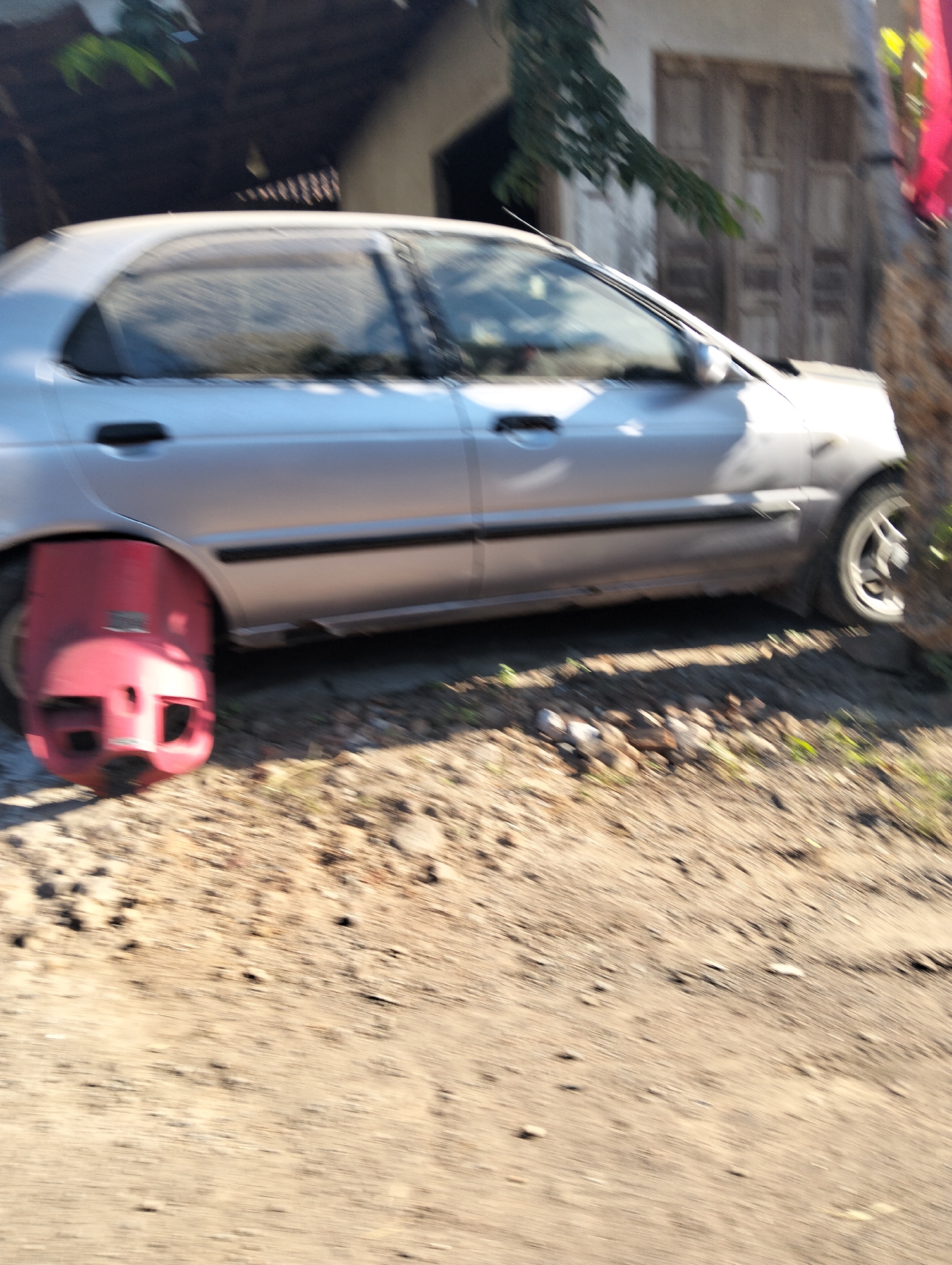 A blurry, medium-shot, outdoor image shows a silver sedan in Kedungringin, Indonesia. The car is parked at an angle, facing left, with its rear wheel on the right side of the frame partially obscured by a tree trunk. The front left tire of the vehicle is missing and has been replaced with a bright red car boot, or wheel clamp, which is prominently displayed. The car is positioned on a dirt road with scattered small rocks and dry vegetation. In the background, there is a weathered building with wooden doors and a dark overhang. Green foliage is visible above and to the left of the car. A sliver of bright pink fabric is also visible on the far right edge of the frame, partially obscuring the tree trunk. The overall image is characterized by motion blur, suggesting it was taken while the vehicle was moving.