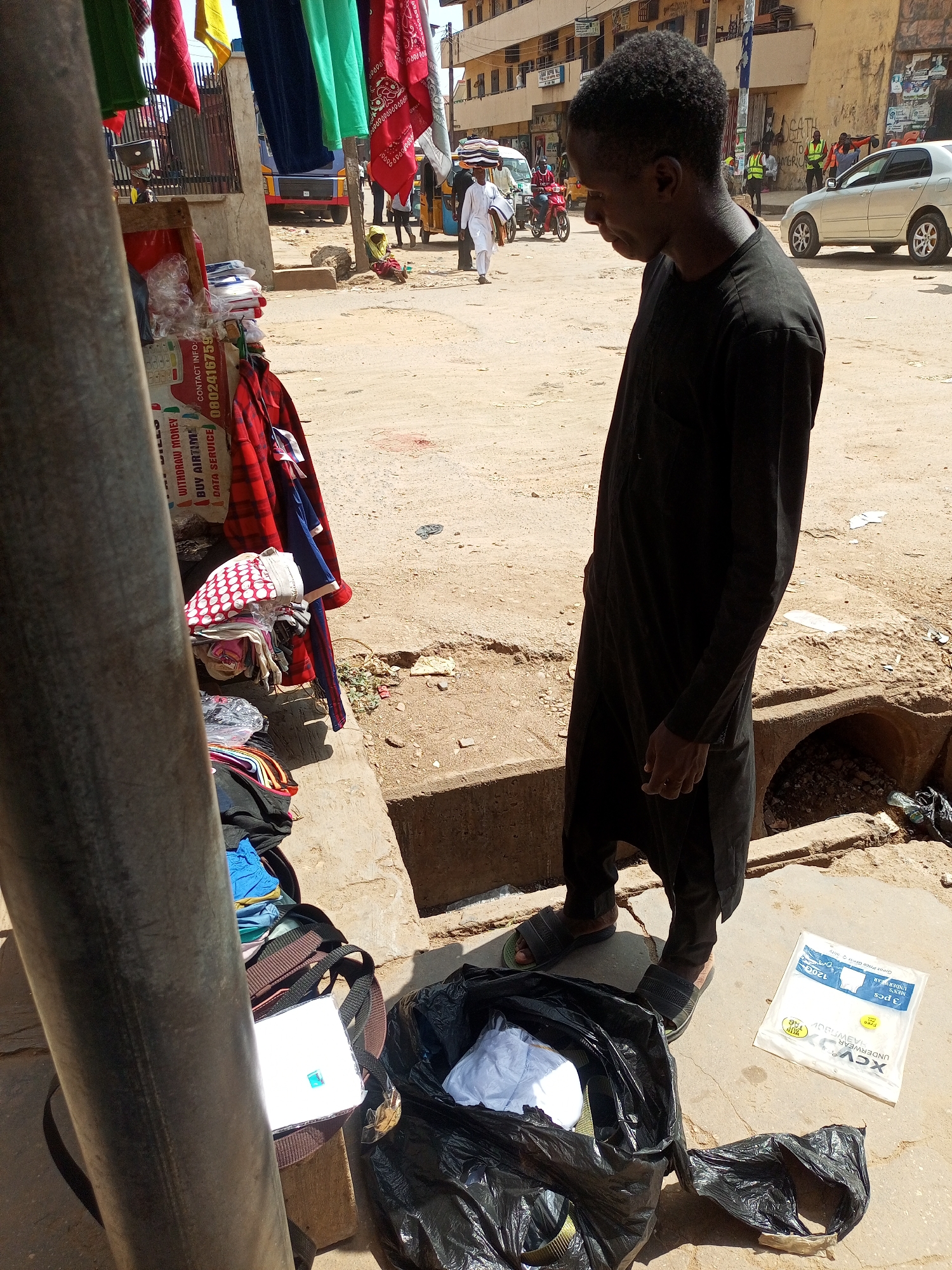 This media file depicts a bustling outdoor street scene in Maiburiji, Nigeria, under bright daylight with harsh shadows indicating sunny weather.
The primary subject is a man standing on the right, dressed in a dark tunic and pants with sandals. He is looking downwards, possibly at the contents of a large black plastic bag at his feet, which contains white items. Next to the bag, a white plastic package of "XCV" brand men's underwear, labeled "3 pcs", "MEN'S UNDERWEAR", and "Great Price," lies on the ground. A brown bag is also visible next to the black plastic bag.
On the left, partially obscured by a metal pole, is a vendor's stall. Colorful fabrics, including red bandanas and various other textiles, hang above. Below, neatly folded clothes and plastic-wrapped items are stacked on a shelf. A prominent sign on the stall advertises services: "WITHDRAW MONEY", "BUY AIRTIME", and "DATA SERVICE", with "CONTACT INFO:" and a phone number "0802416759" clearly visible.
The background reveals a lively street environment. The ground is a mix of dirt and concrete, with a concrete drainage ditch running along the side. Various people are visible further back, including one person carrying goods on their head and others walking. A gold sedan and a blue truck are parked or moving in the distance, and individuals wearing high-visibility vests can be seen. Multi-story buildings line the street, suggesting a commercial or residential area. The overall scene portrays everyday market activity.