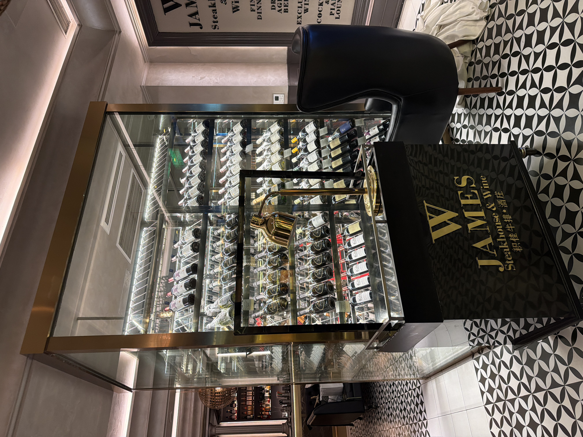 The photograph presents a detailed view of a high-end steakhouse and wine establishment, likely located in Qinhuai District, China. A large, glass-encased wine cellar dominates the foreground, filled with numerous bottles of wine arranged on illuminated racks. A golden service faucet extends from the cellar, adding a touch of opulence.
Next to the cellar stands a black, stylized sign displaying the name "JAMES Steakhouse & Wine," with the name "JAMES" in large gold letters and the Chinese translation "积佳牛排・酒庄" underneath. A black leather chair sits beside the sign, suggesting a comfortable and upscale ambiance.
The floor is covered in a patterned black and white tile, which contrasts with the neutral-colored walls and ceiling. Behind the wine cellar, a glimpse of the restaurant's interior is visible, including stairs, a decorative light fixture, and a seating area. A menu board listing various dining options, including "WINE," "COCK," and "PIANO LOUNGE," is affixed to the wall.