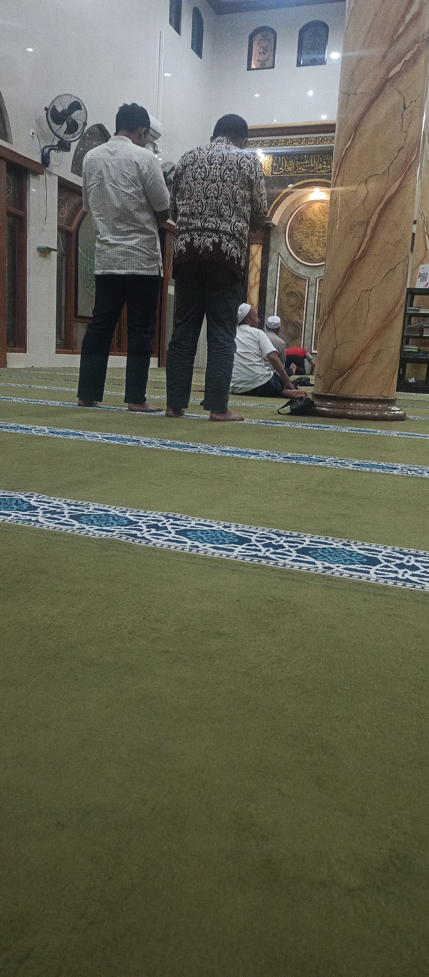 In a mosque in Arjasa, Indonesia, three men are engaged in prayer. Two men stand facing a decorative mihrab on the far wall, their backs to the camera. The man on the left wears a white patterned shirt and dark trousers. The man on the right wears a dark patterned shirt and dark trousers. A third man sits on the green carpeted floor in front of them, facing the mihrab. He wears a white cap and white shirt, with dark trousers. Another man, partially obscured, is in a kneeling prayer position behind him. The mosque interior features a large decorative pillar on the right, intricate calligraphy on the wall, and patterned borders on the carpet.