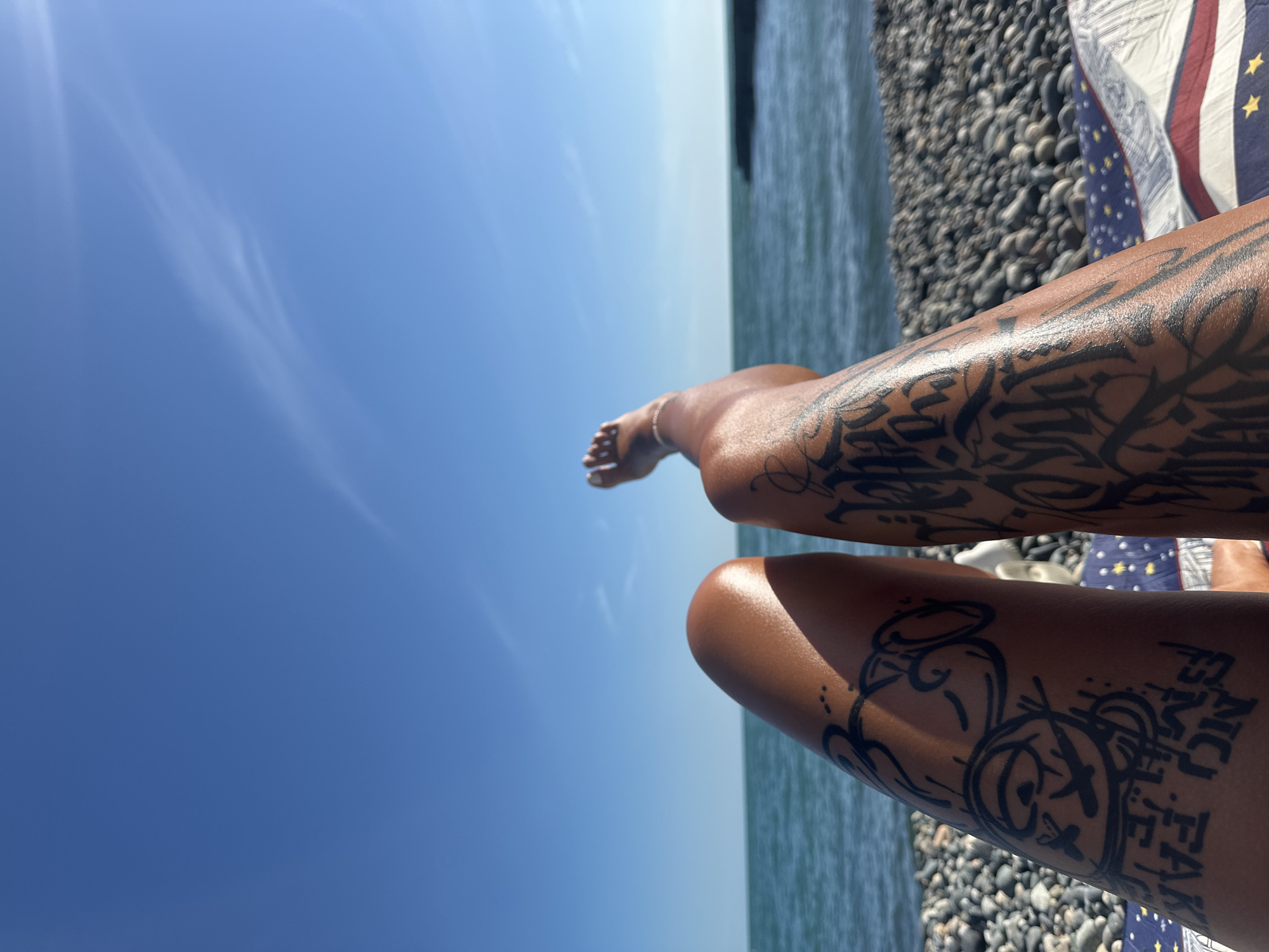 The image depicts a first-person perspective of a person lying on a pebbled beach with their legs extended towards the sea. The sky is a clear, bright blue with a few wispy clouds. The sea is a deep blue-green and appears calm. The person's legs, covered in black tattoos, are the primary focus in the foreground. One leg has a tattoo resembling a cartoon character with an eye, and the other has script-style lettering. The person is wearing a light-colored anklet. A patterned towel or blanket lies beneath the legs. The scene is likely located on a beach in the city of New Athos, Abkhazia, characterized by its pebbled shores and clear waters.
