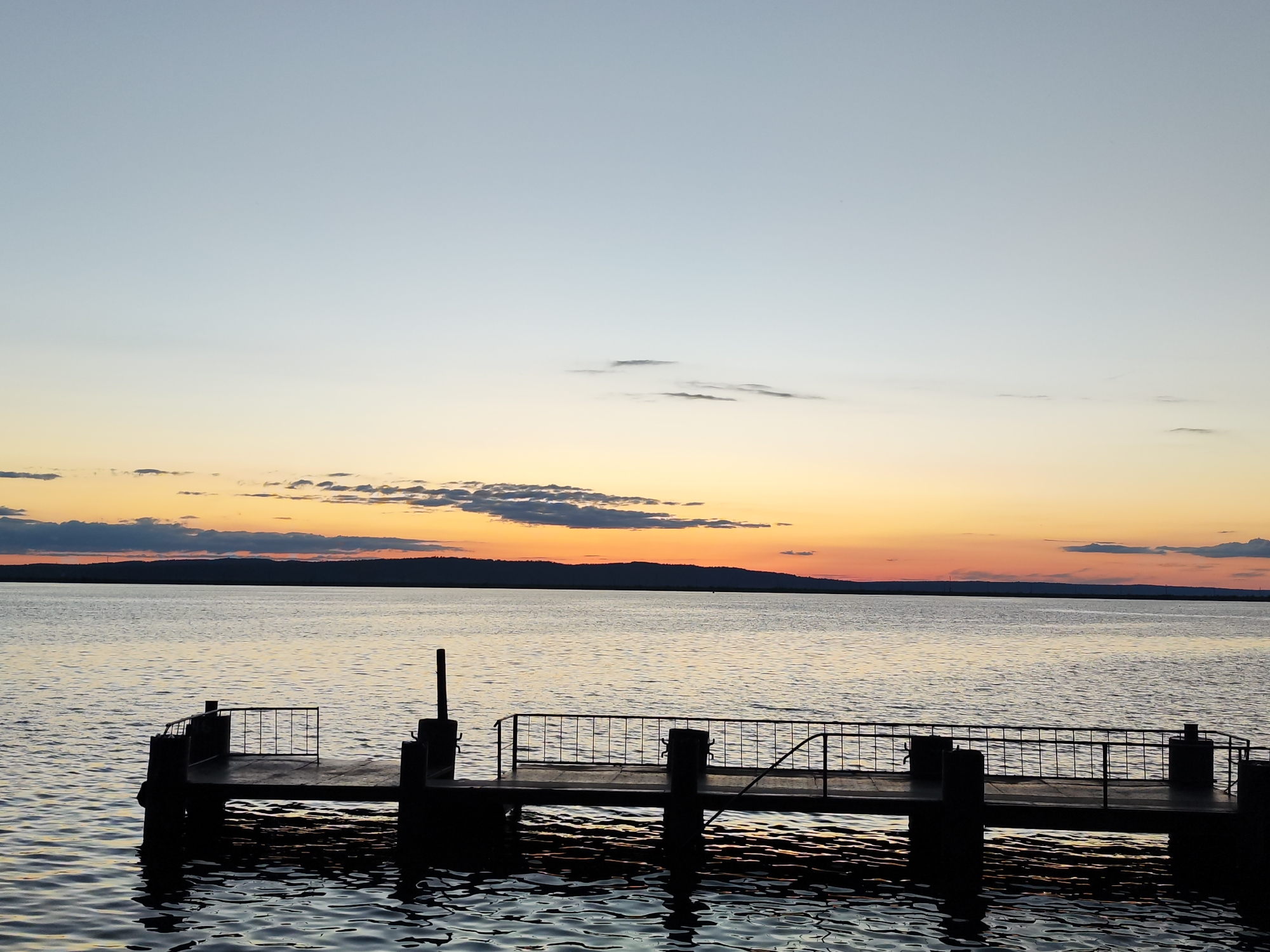 The image presents a tranquil landscape at either sunrise or sunset over a large body of water, likely the Votkinsk Reservoir on the Kama River near Chaikovsky, Russia. In the foreground, the dark silhouette of a pier or dock extends into the water from the left. The pier features robust posts and a metal railing that casts reflections onto the gently rippling water below.
The expansive body of water fills the middle ground, its surface reflecting the subtle hues of the sky. A distant, dark landmass, appearing as a low treeline or hill range, stretches across the horizon, creating a stark silhouette against the brightly colored sky.
The sky is a prominent feature, displaying a beautiful gradient of colors typical of twilight. The lower sky, closest to the horizon, glows with warm shades of orange, yellow, and soft pink. This transitions upwards into cooler tones of light blue and grey. Several scattered, wispy clouds are visible, particularly along the horizon, catching the warm light, while smaller, darker clouds are dispersed higher in the cooler sky.
The scene conveys a sense of peace and serenity. The calm water and the soft, low light suggest quiet weather conditions. There are no people visible, and no text can be discerned in the image.